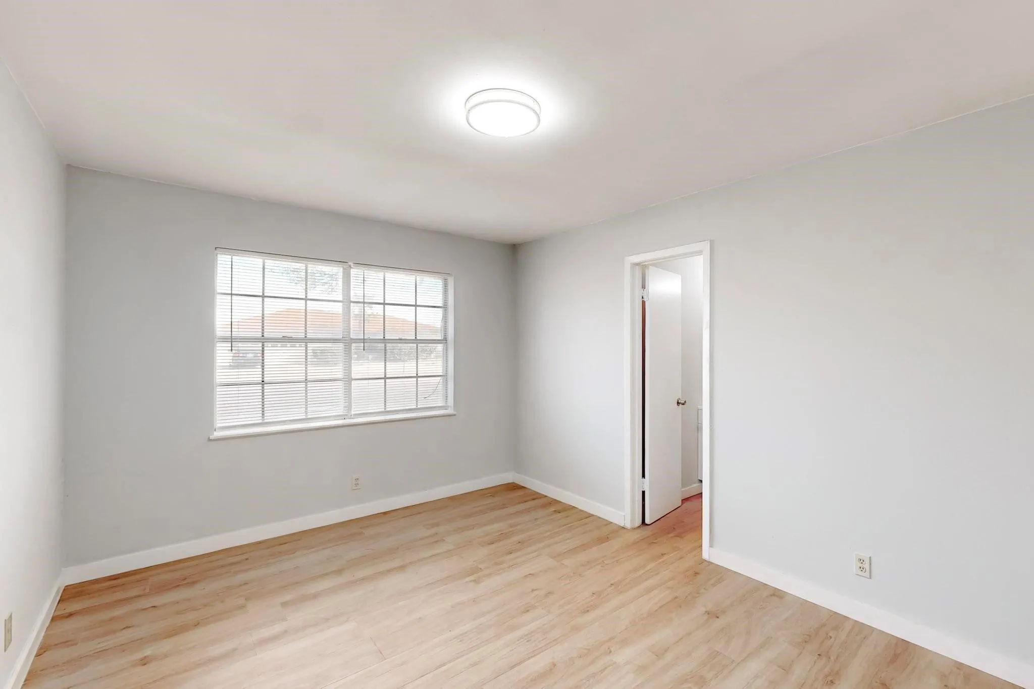 Wood-finish flooring with light-toned walls, featuring a ceiling-mounted light fixture and a window with horizontal blinds