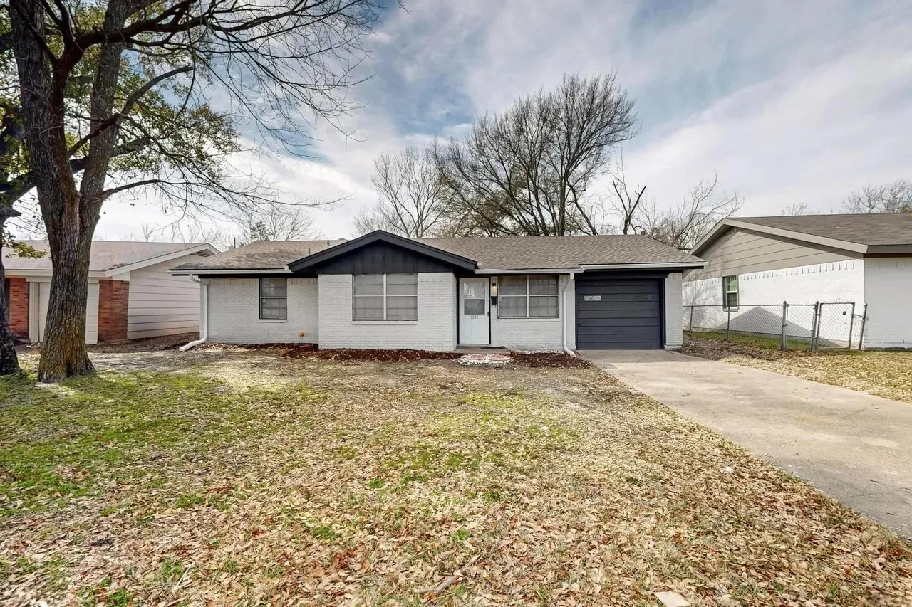 White brick exterior, black trim accents, attached garage, paved driveway, and established trees