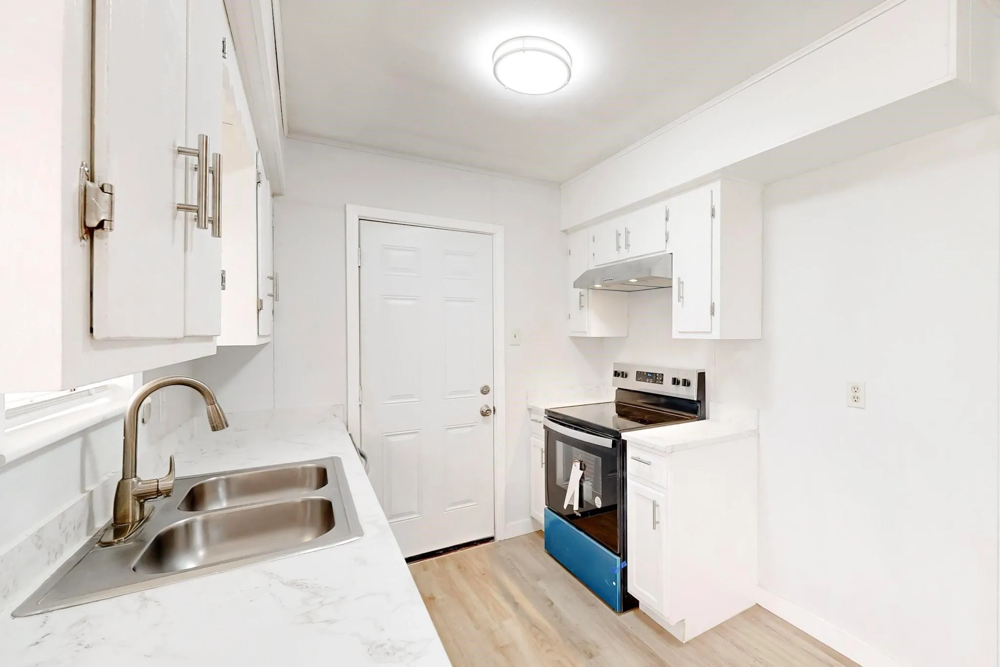 White cabinetry, stainless steel double basin sink, modern faucet, wood-finish flooring, and a built-in electric range with exhaust hood