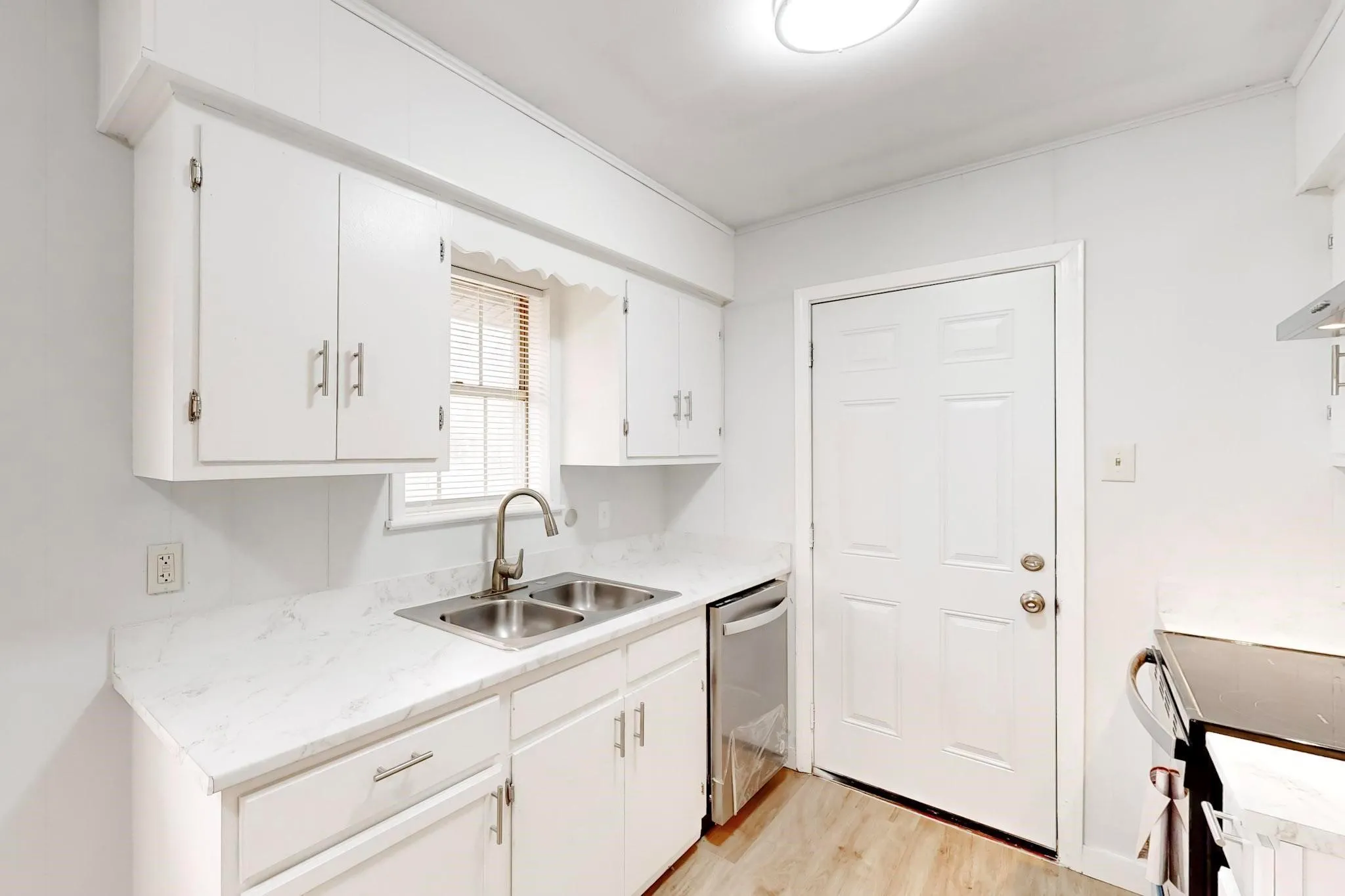 White cabinetry with brushed nickel hardware, stainless steel double basin sink, light-toned countertops, wood-finish flooring, and a stainless steel dishwasher