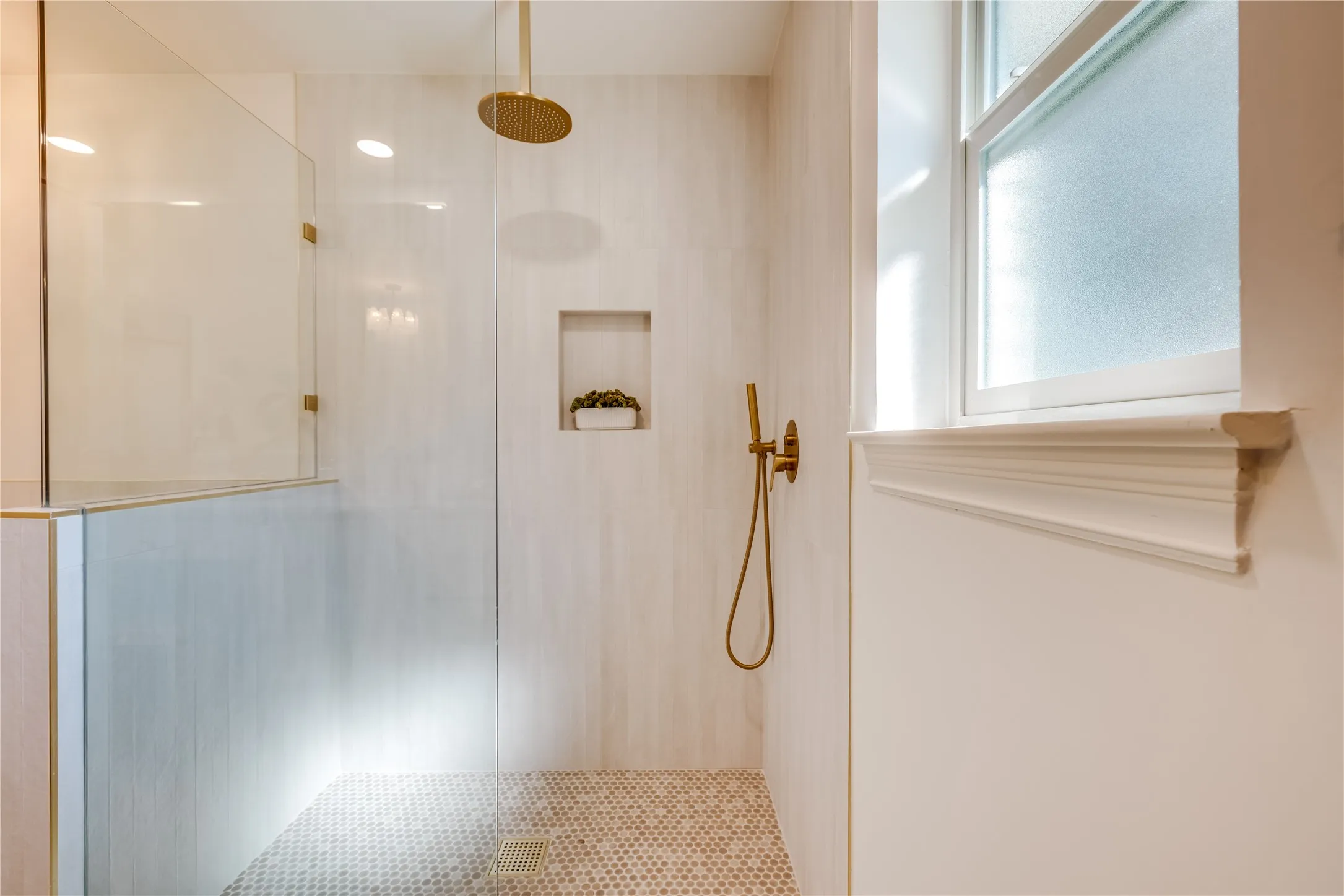 Walk-in shower featuring a frameless glass enclosure, tiled walls, hexagonal mosaic floor, recessed niche, and contemporary gold-finish fixtures