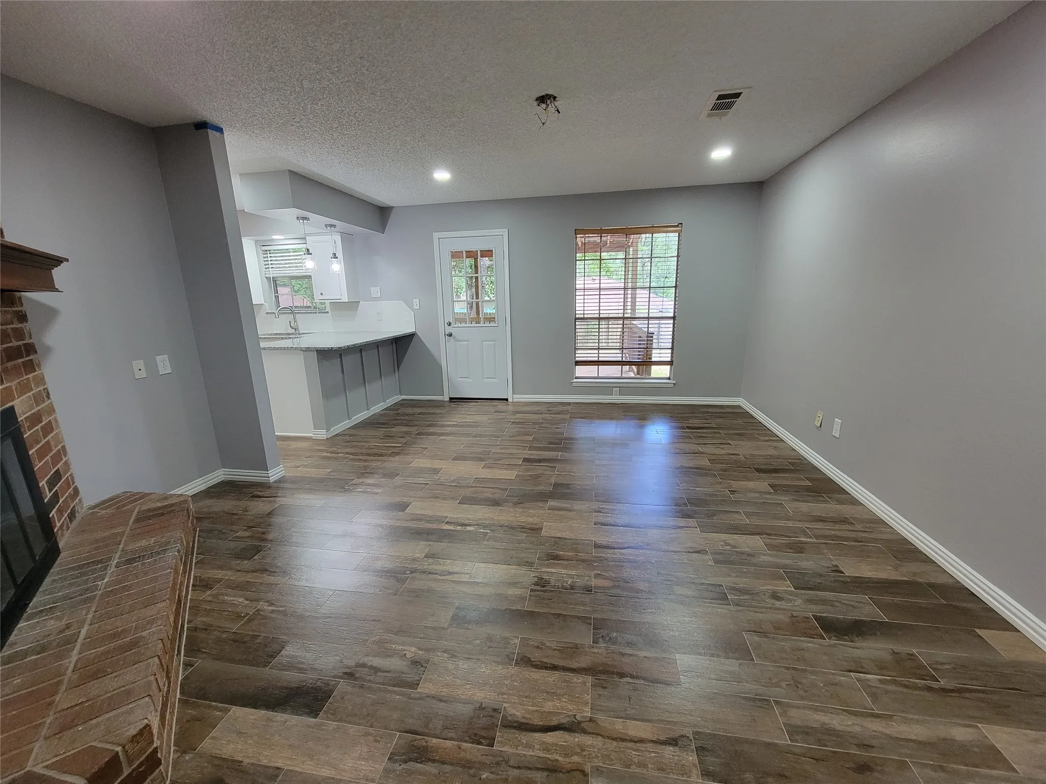 Wood-finish tile flooring throughout, recessed ceiling lighting, a brick fireplace, and a white kitchen with a breakfast bar