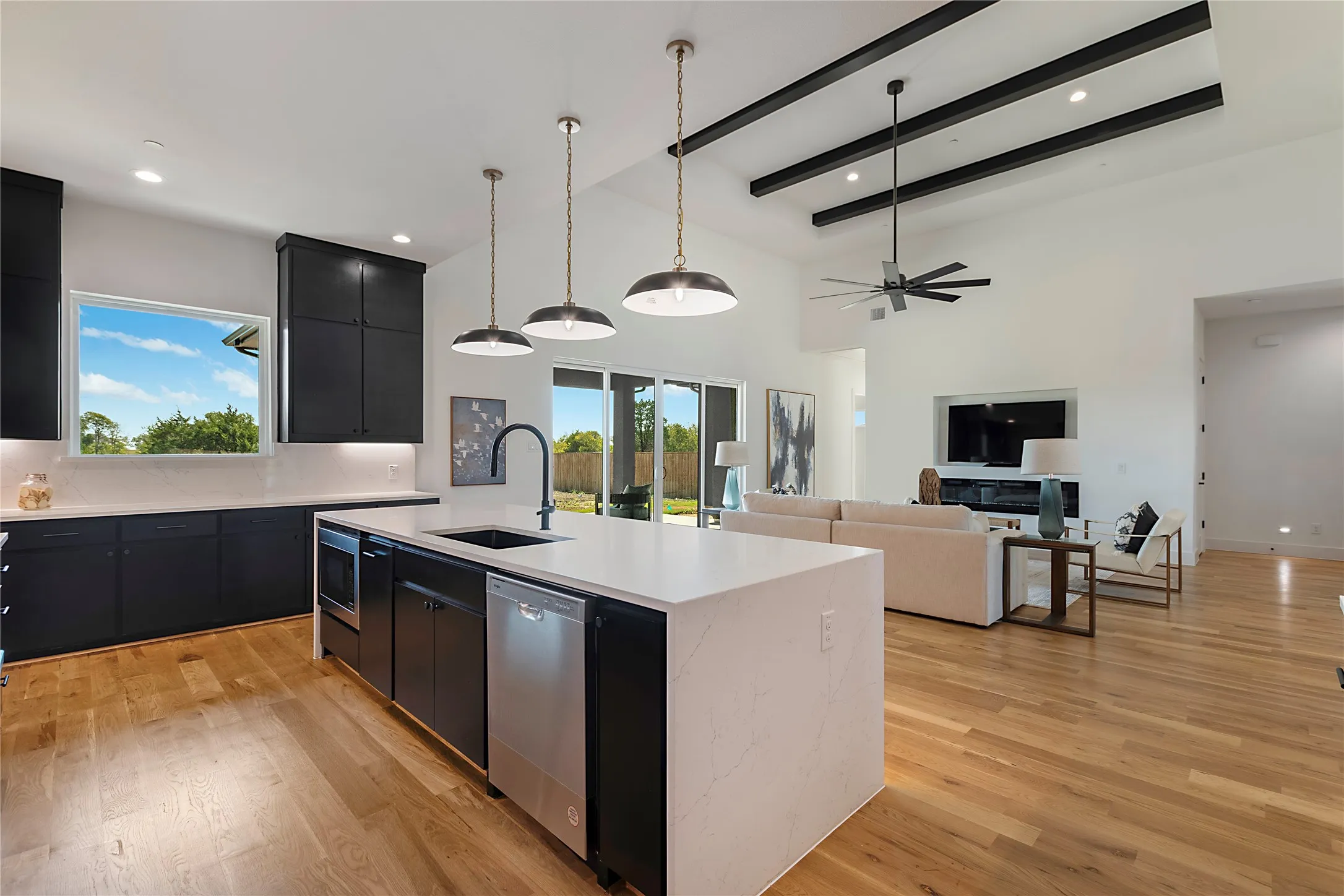 Open-concept kitchen and living area showcasing white quartz waterfall-edge island with integrated cooktop and dishwasher, black cabinetry, stainless steel appliances, pendant lighting, exposed beam ceiling detail, wood-finish flooring throughout, and abundant natural light from multiple windows and glass doors