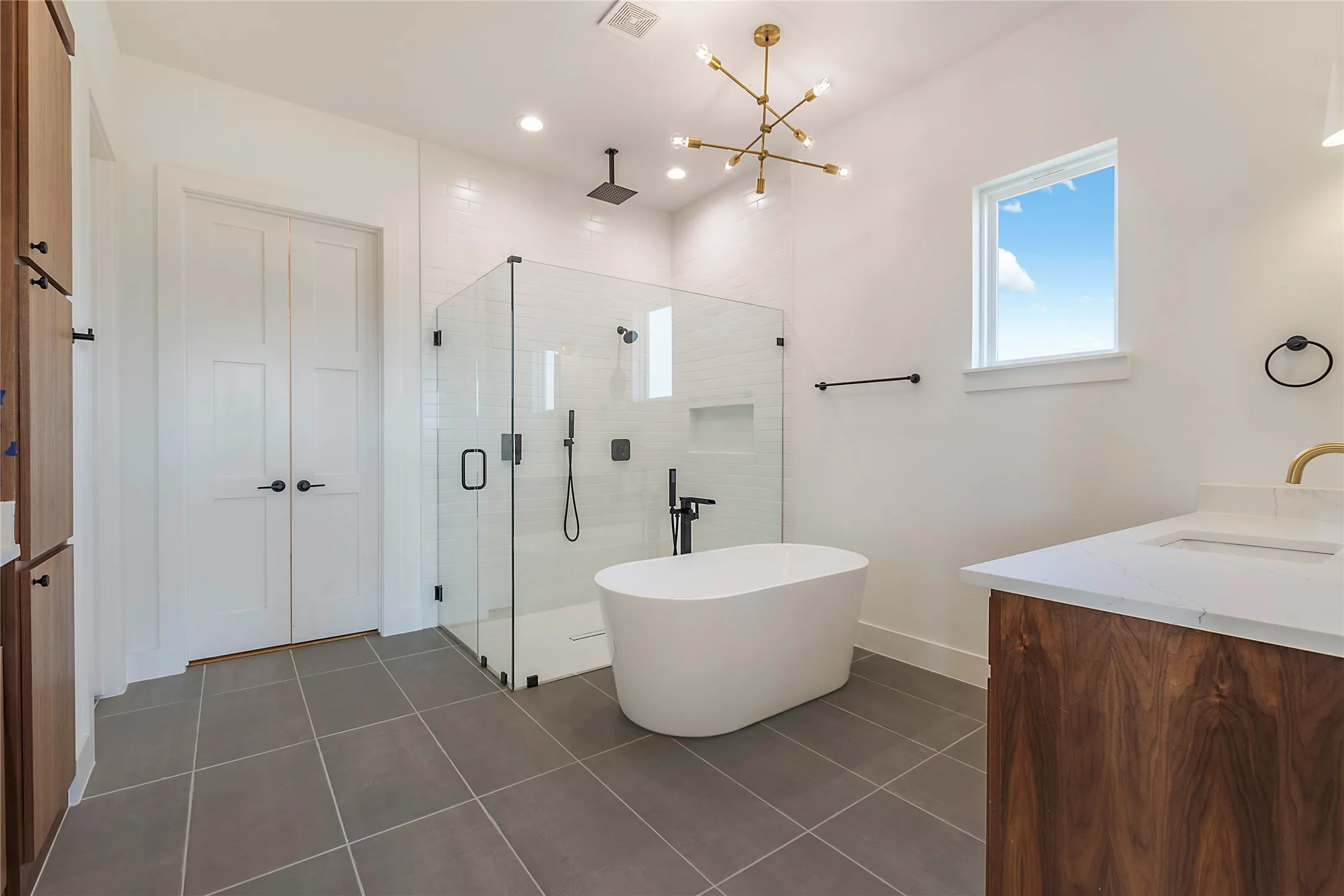 Spa-inspired primary bathroom featuring a freestanding soaking tub, dual vanities with wood-finish cabinetry, frameless glass shower enclosure with rainfall showerhead, radiant heated flooring, and natural light from a skylight