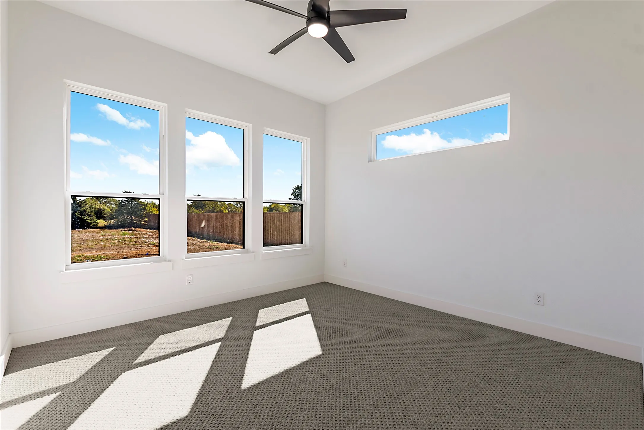 White walls, gray patterned carpeting, three tall windows, one horizontal transom window, and a ceiling fan with a black finish