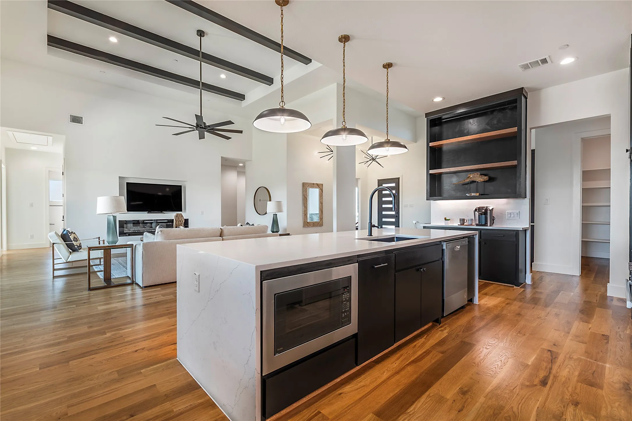 Expansive kitchen island with a built-in microwave and quartz countertop, wood-finish flooring, vaulted ceilings with dark wood beams, and a built-in shelving unit