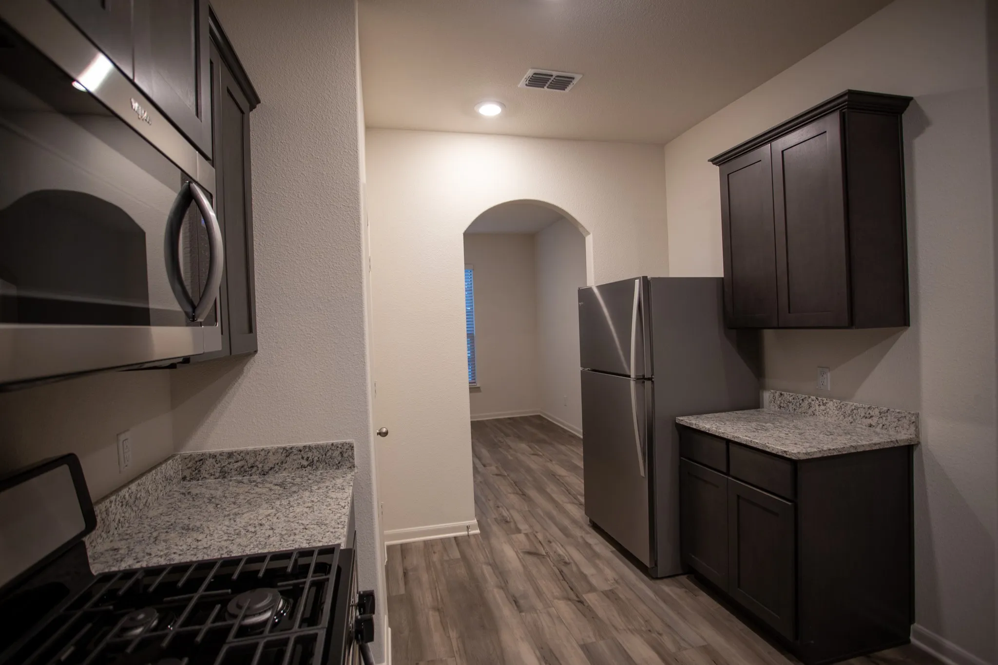 Kitchen featuring stainless steel appliances, dark wood-finish cabinetry, light-toned countertops, wood-finish flooring, and an arched doorway