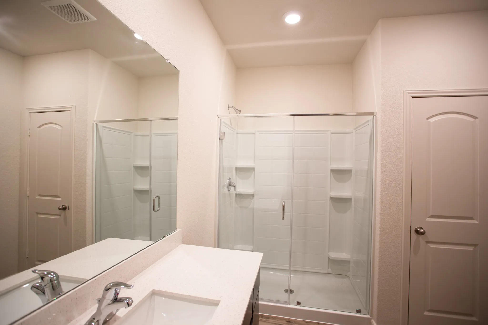 Vanity with integrated sink and chrome faucet, large frameless mirror, and glass-enclosed shower with tiled walls and built-in shelving