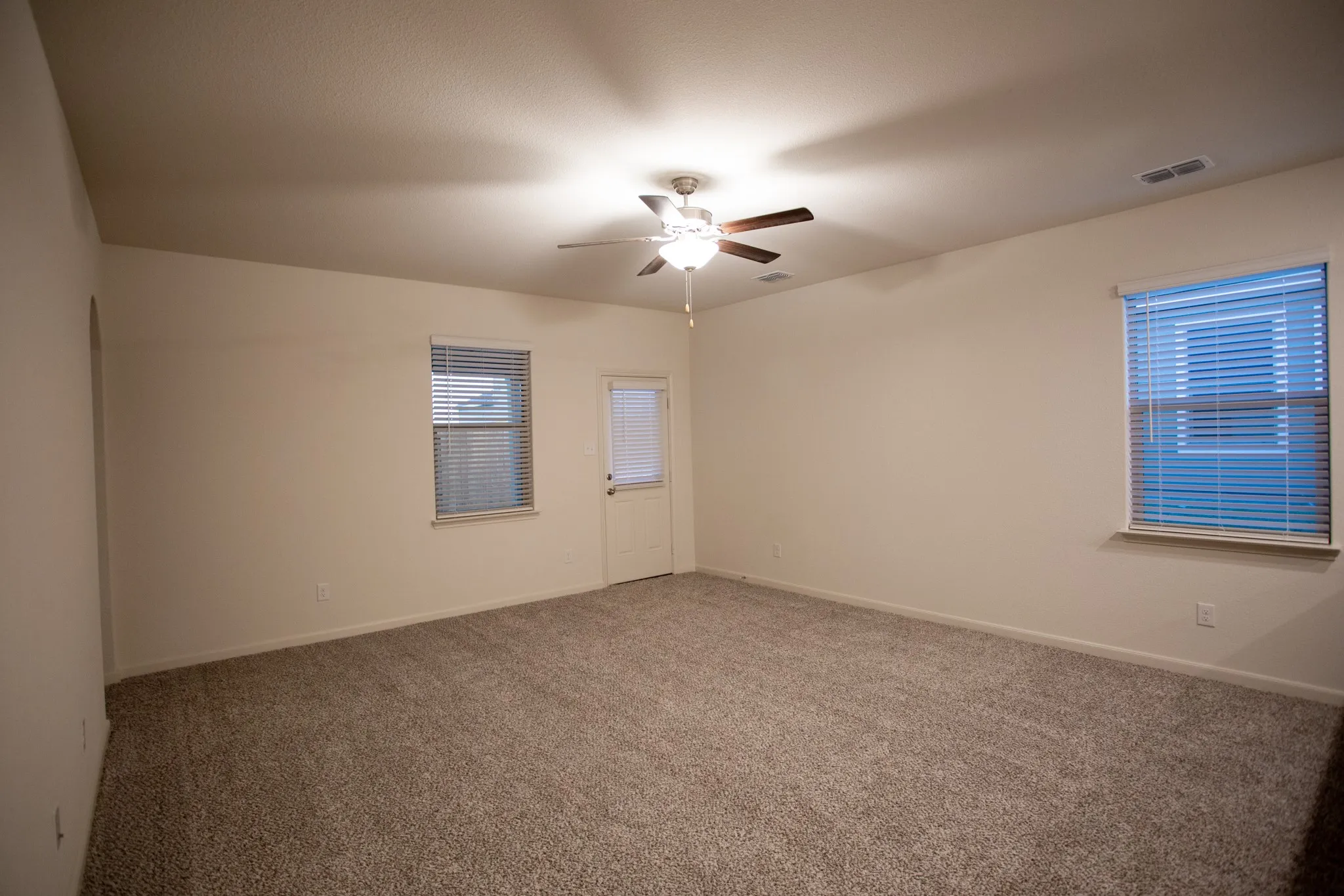 Spacious room with neutral carpet, two windows with blinds, a ceiling fan with integrated lighting, and a white door with an integrated blind