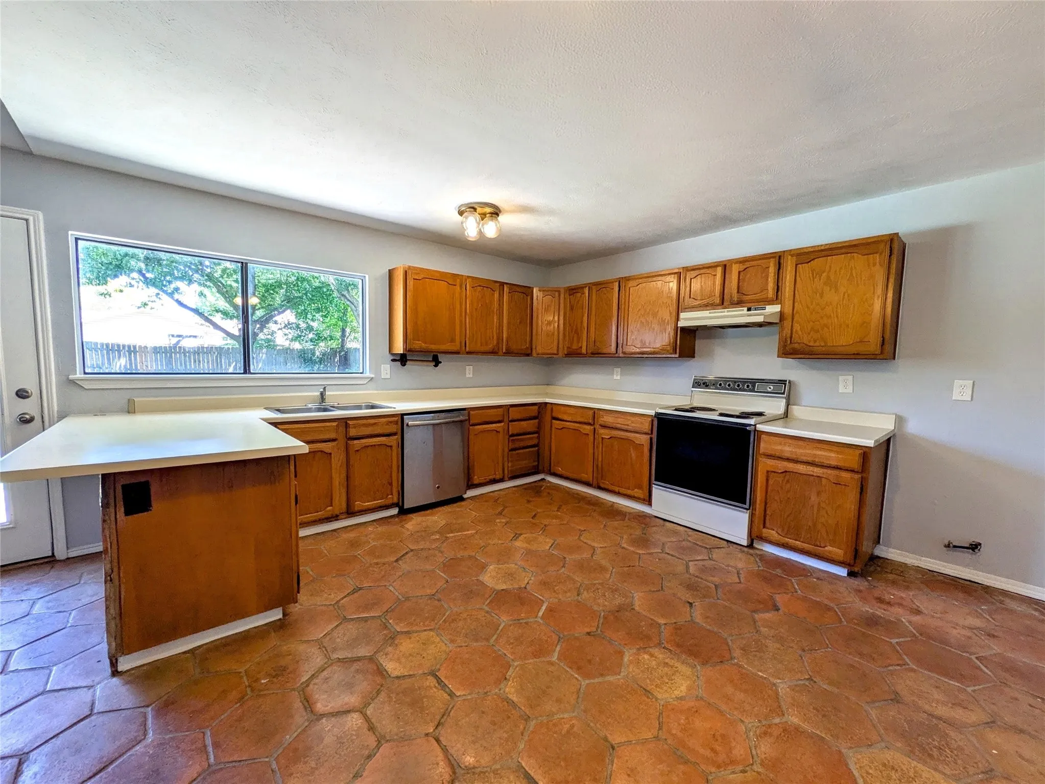 Kitchen featuring hexagonal tile flooring, wood cabinetry, a stainless steel dishwasher, a window with exterior views, and a ceiling-mounted light fixture
