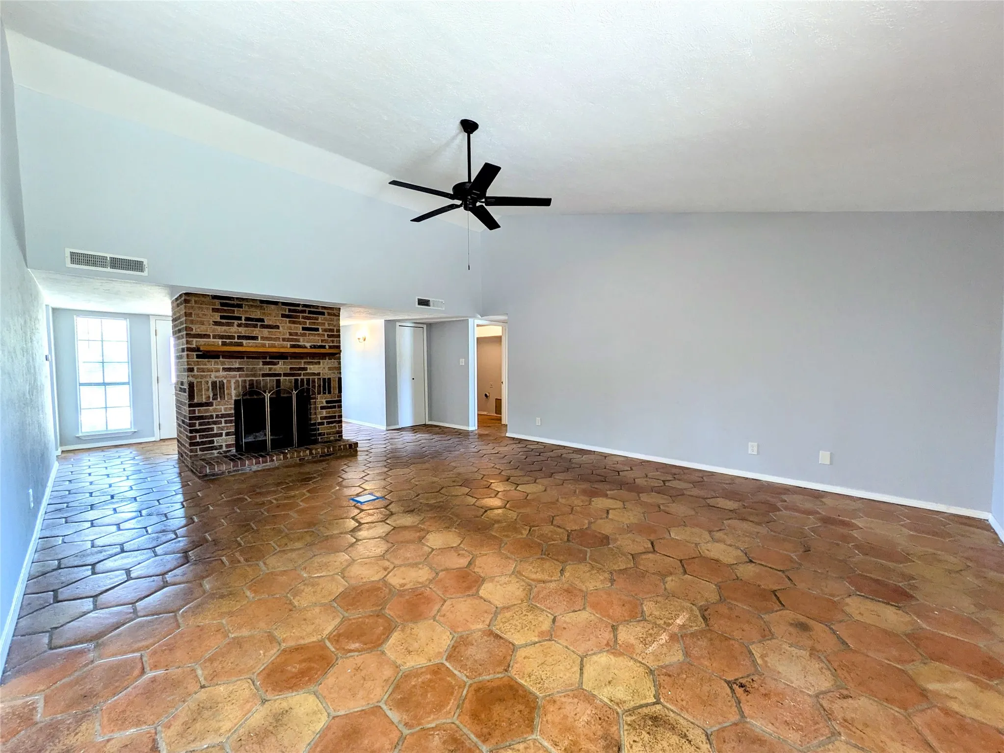Spacious room featuring hexagonal terracotta-style tile flooring, a brick fireplace, a vaulted ceiling, a ceiling fan, and a double window