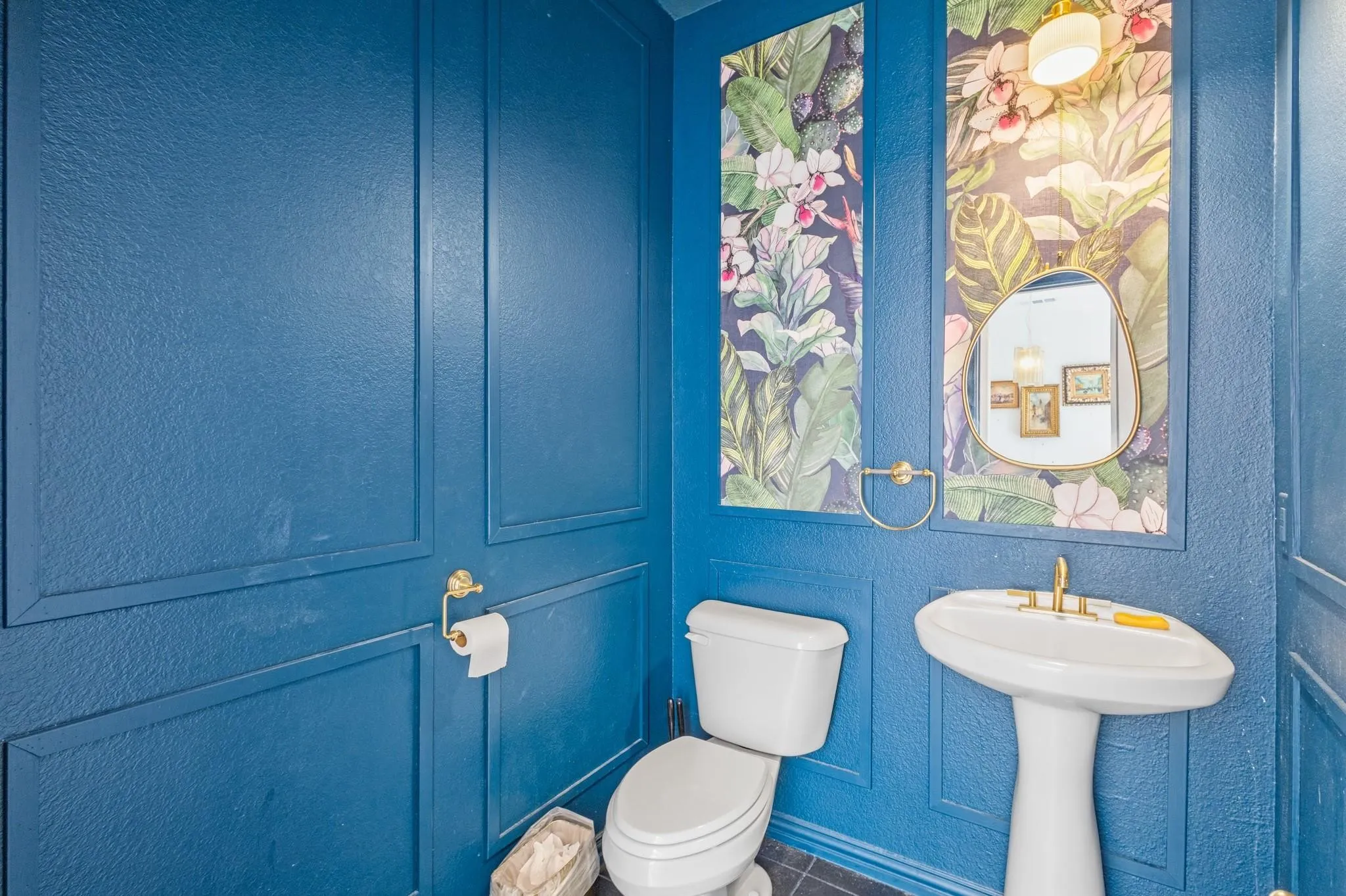 Pedestal sink with brass-finish faucet, wall-mounted mirror, accent wall with tropical-patterned panels, and paneled wall finishes