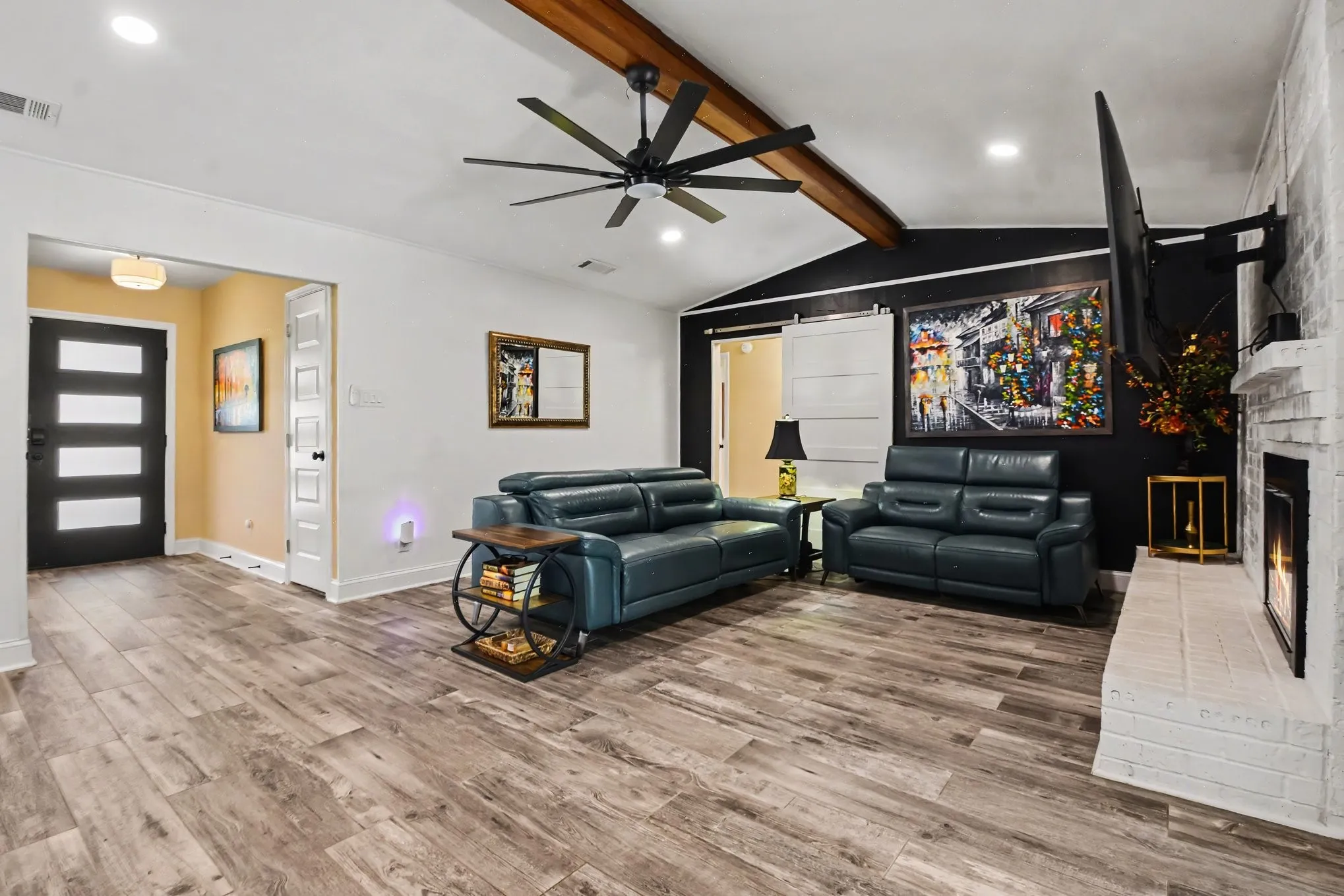 Vaulted ceiling with wood beam, wood-finish flooring, white brick fireplace, recessed lighting, and a modern ceiling fan
