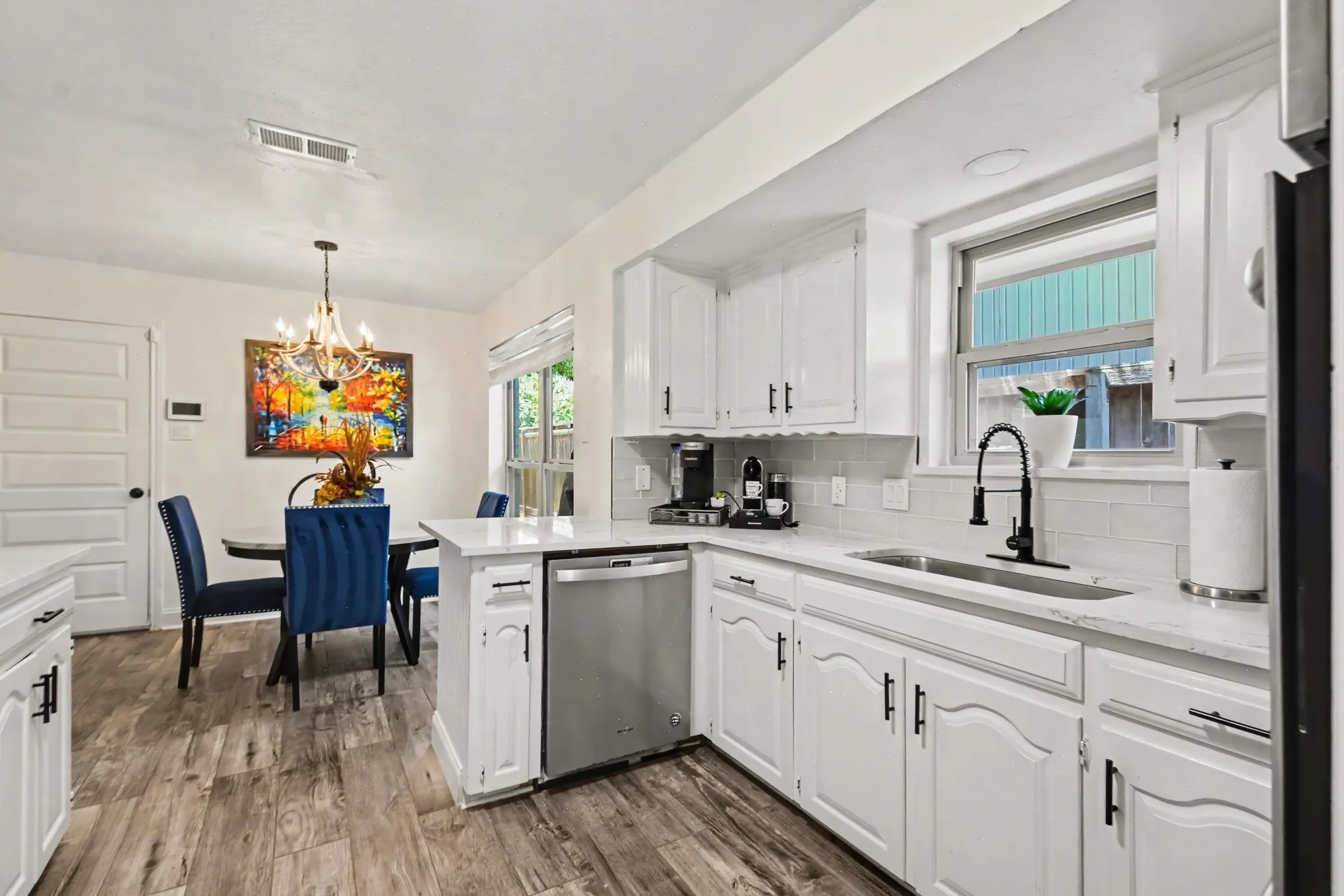 White kitchen cabinetry, stainless steel dishwasher, wood-finish flooring, undermount sink with a black gooseneck faucet, and light-toned backsplash