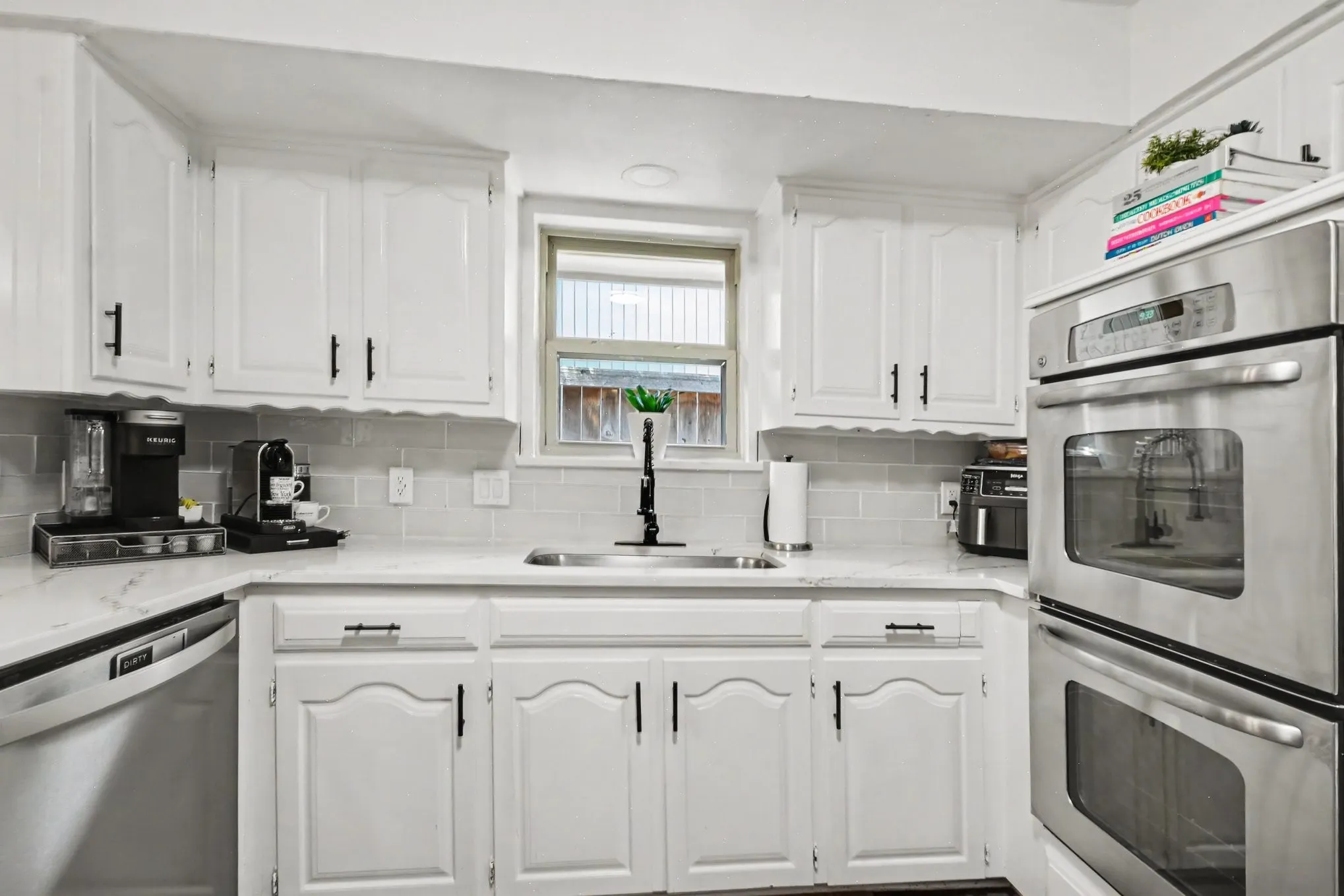 White cabinetry with black hardware, light-toned countertops, subway tile backsplash, stainless steel double ovens, and an undermount sink with a black gooseneck faucet