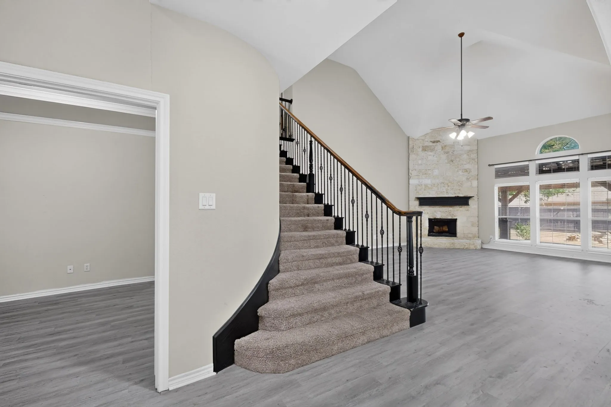 Curved staircase with black wrought iron spindles, stone fireplace, vaulted ceiling, large windows, and wood-finish flooring