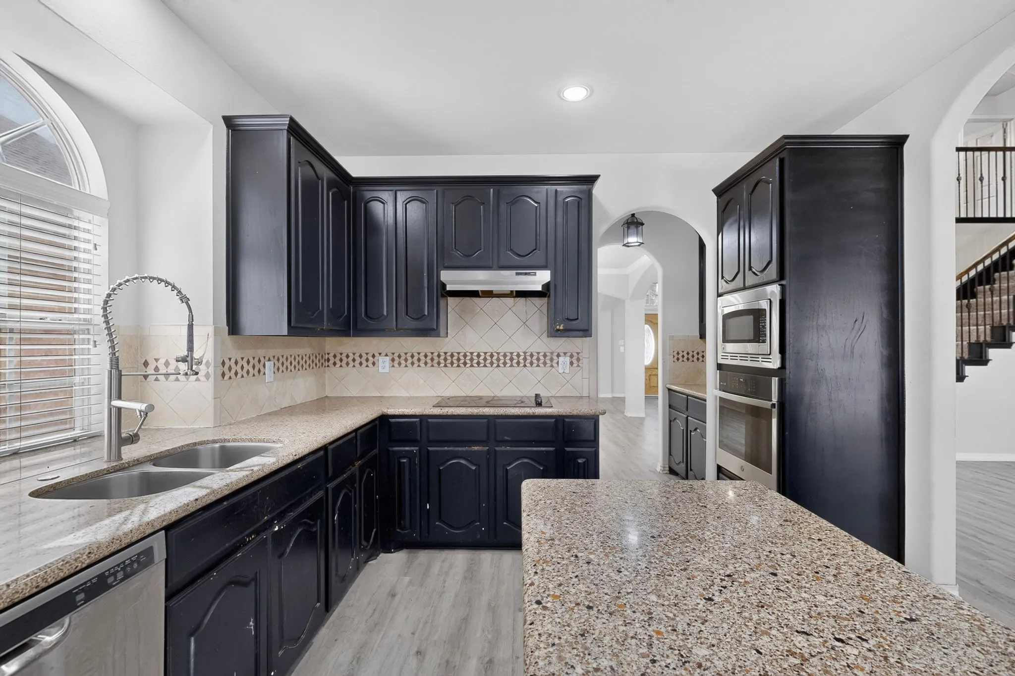 Kitchen featuring dark cabinetry, light-toned countertops, a stainless steel double sink with a pull-down faucet, integrated stainless steel appliances, and wood-finish flooring