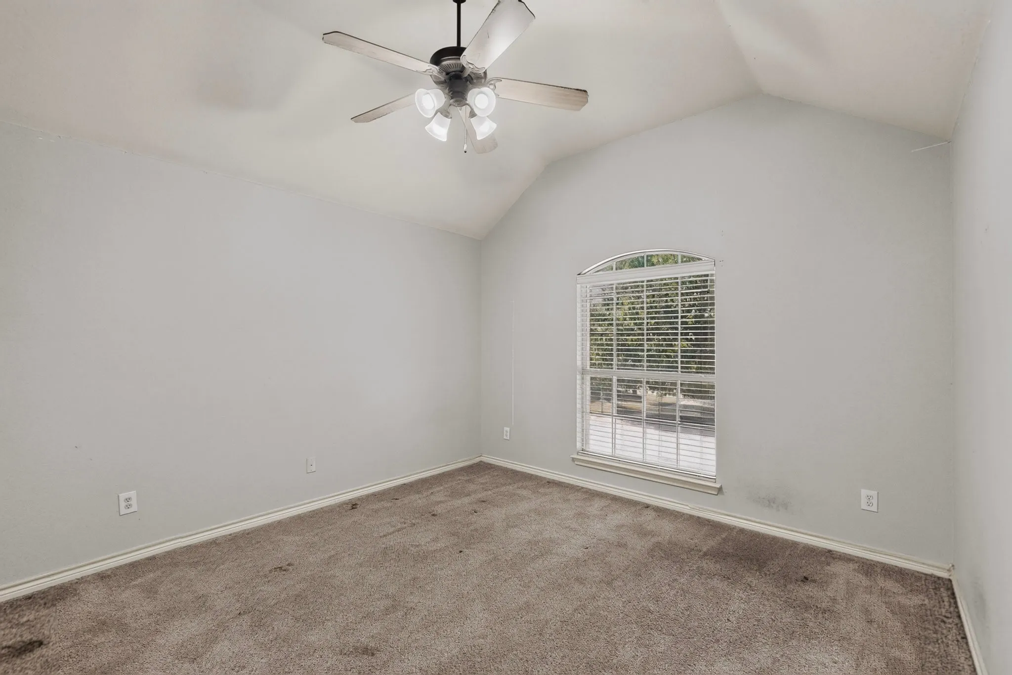 Spacious bedroom with vaulted ceiling, ceiling fan with integrated lighting, neutral carpet flooring, and large window with plantation shutters providing natural light