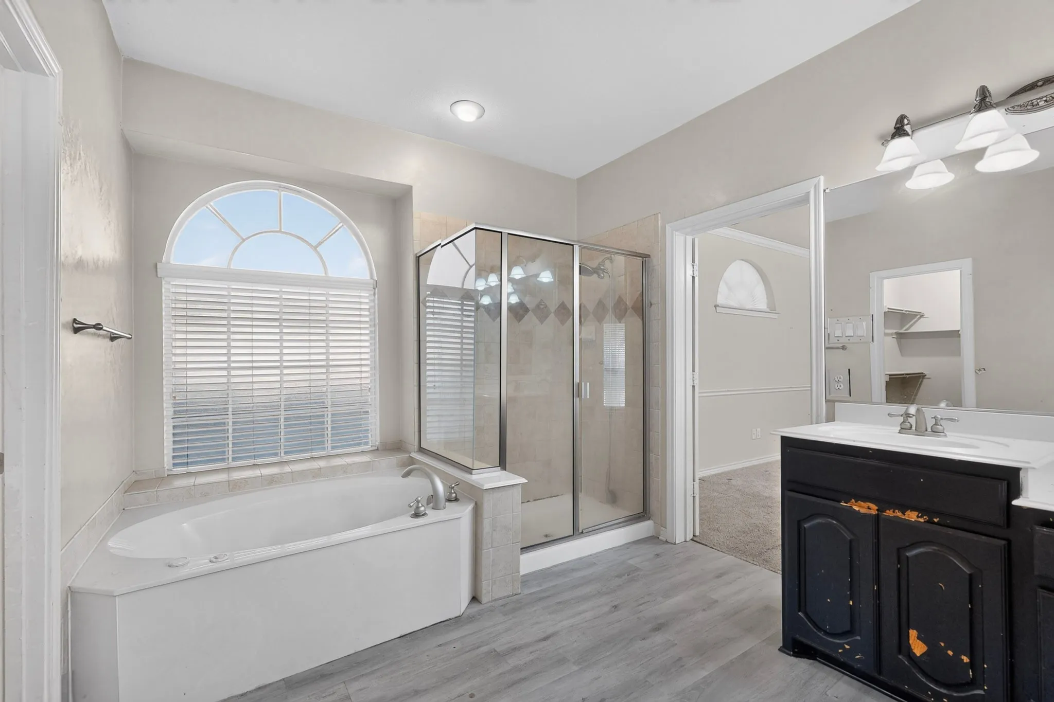 Arch-top window with blinds, built-in bathtub, glass-enclosed shower, wood-finish flooring, and double vanity with integrated sinks