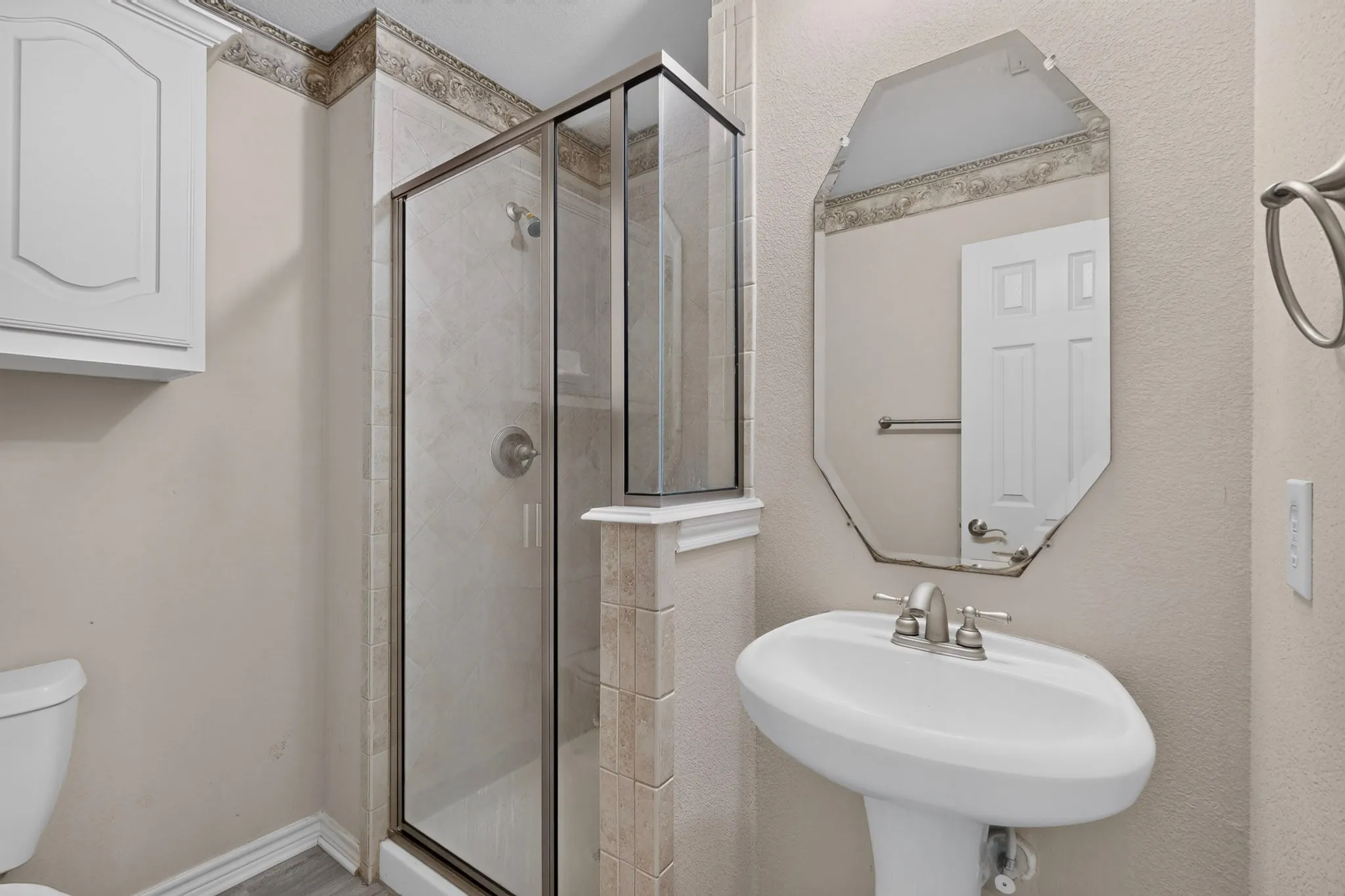 Glass-enclosed shower with tiled walls, pedestal sink with brushed nickel faucet, and a wall-mounted storage cabinet