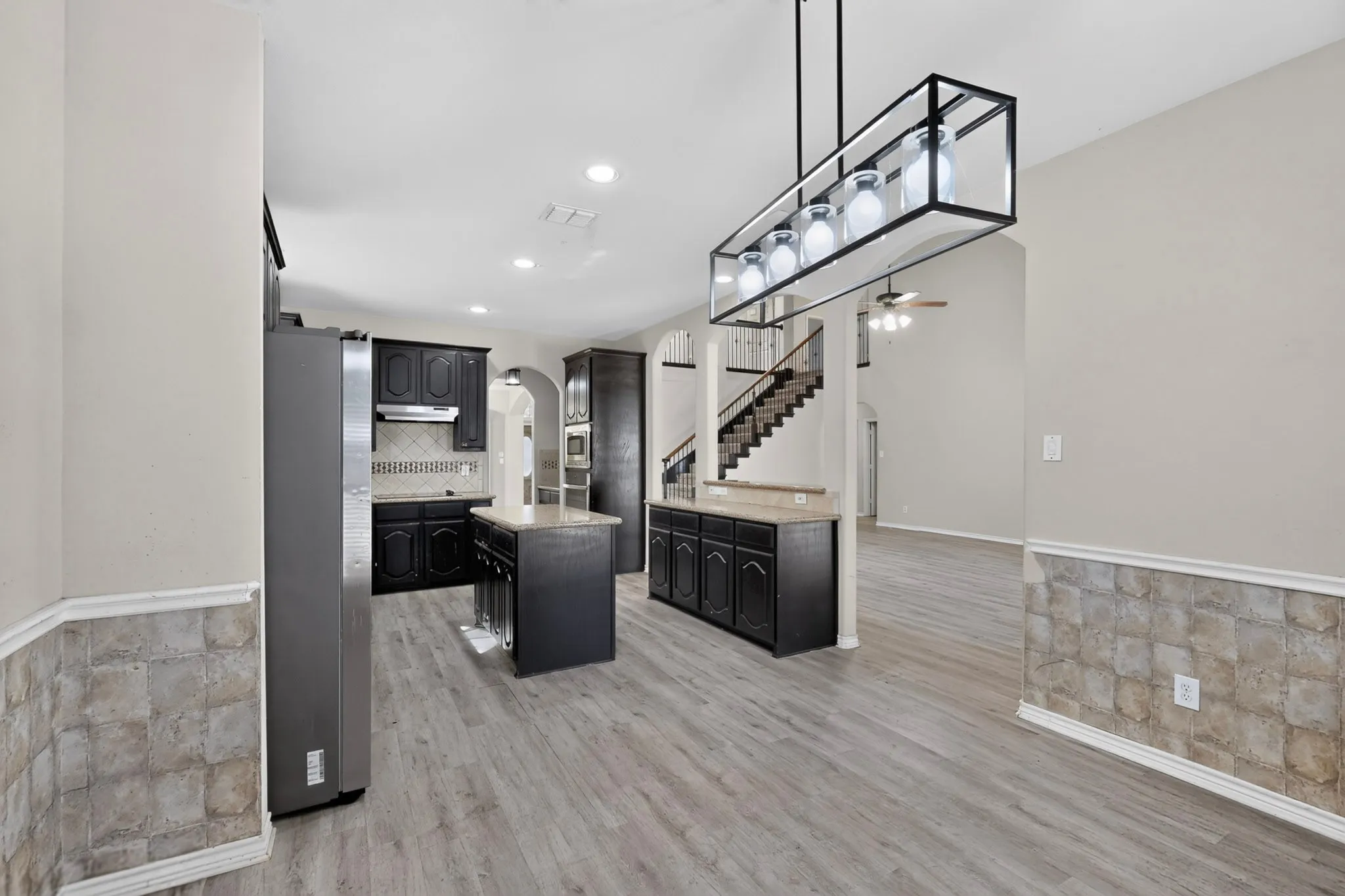 Wood-finish flooring throughout, kitchen with dark cabinetry and light countertops, center island, contemporary linear chandelier, and staircase with dark railings