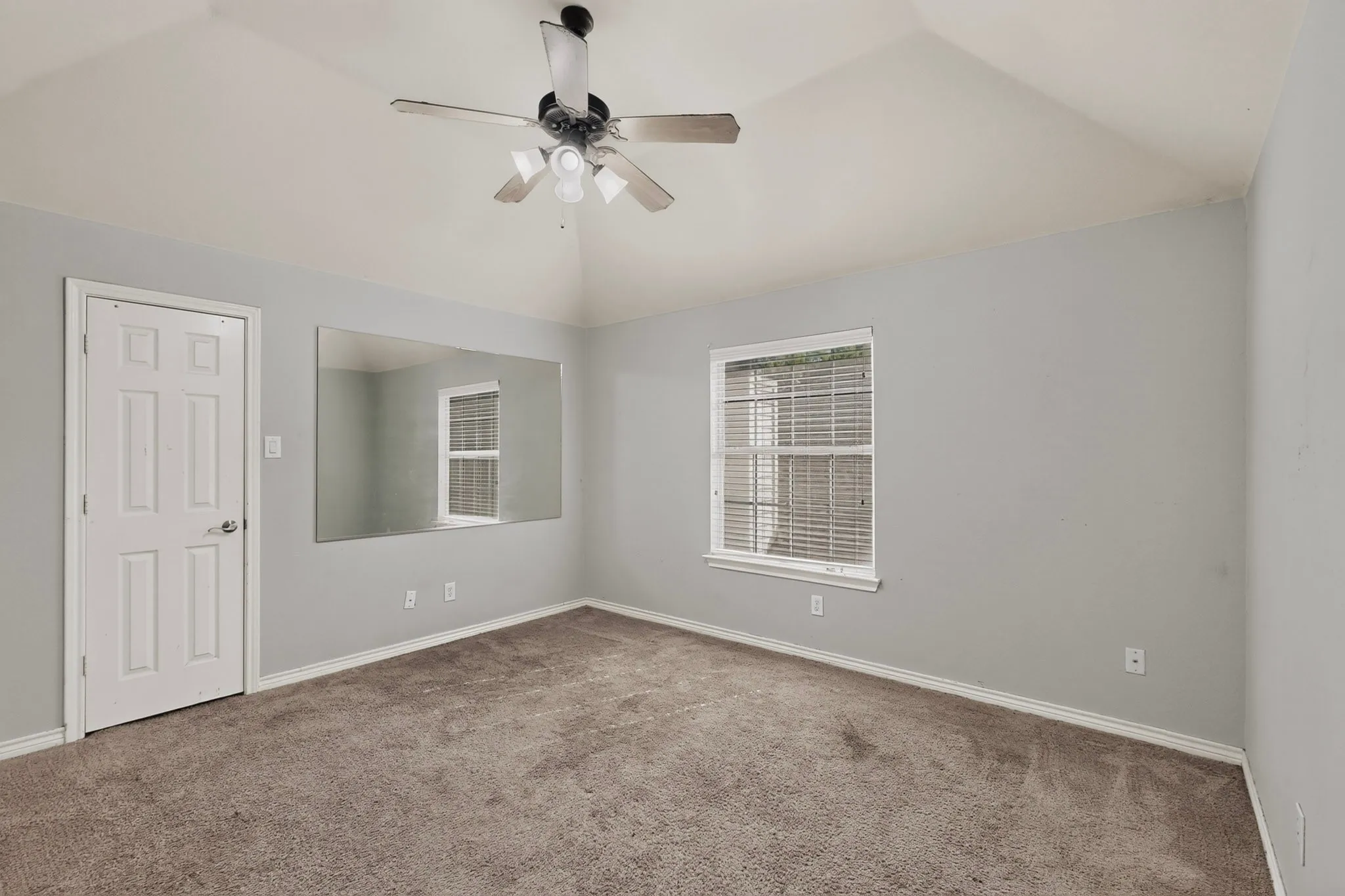Vaulted ceiling with ceiling fan, large wall-mounted mirror, single window with blinds, light gray wall paint, and carpeted flooring