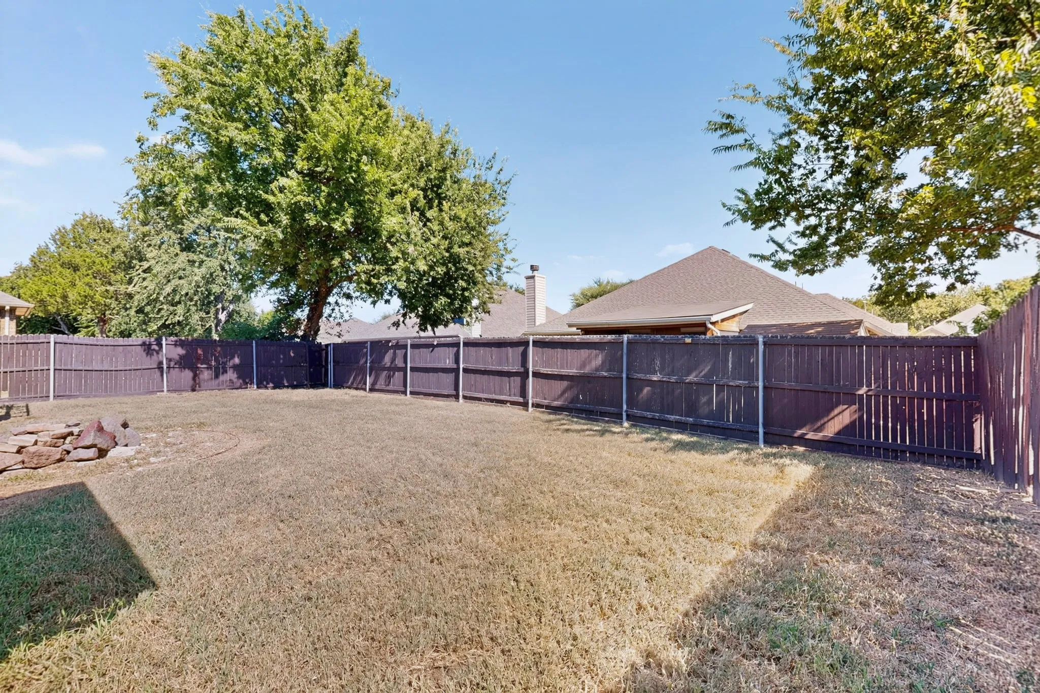 Expansive backyard featuring a privacy fence, mature trees, and a stone-lined fire pit