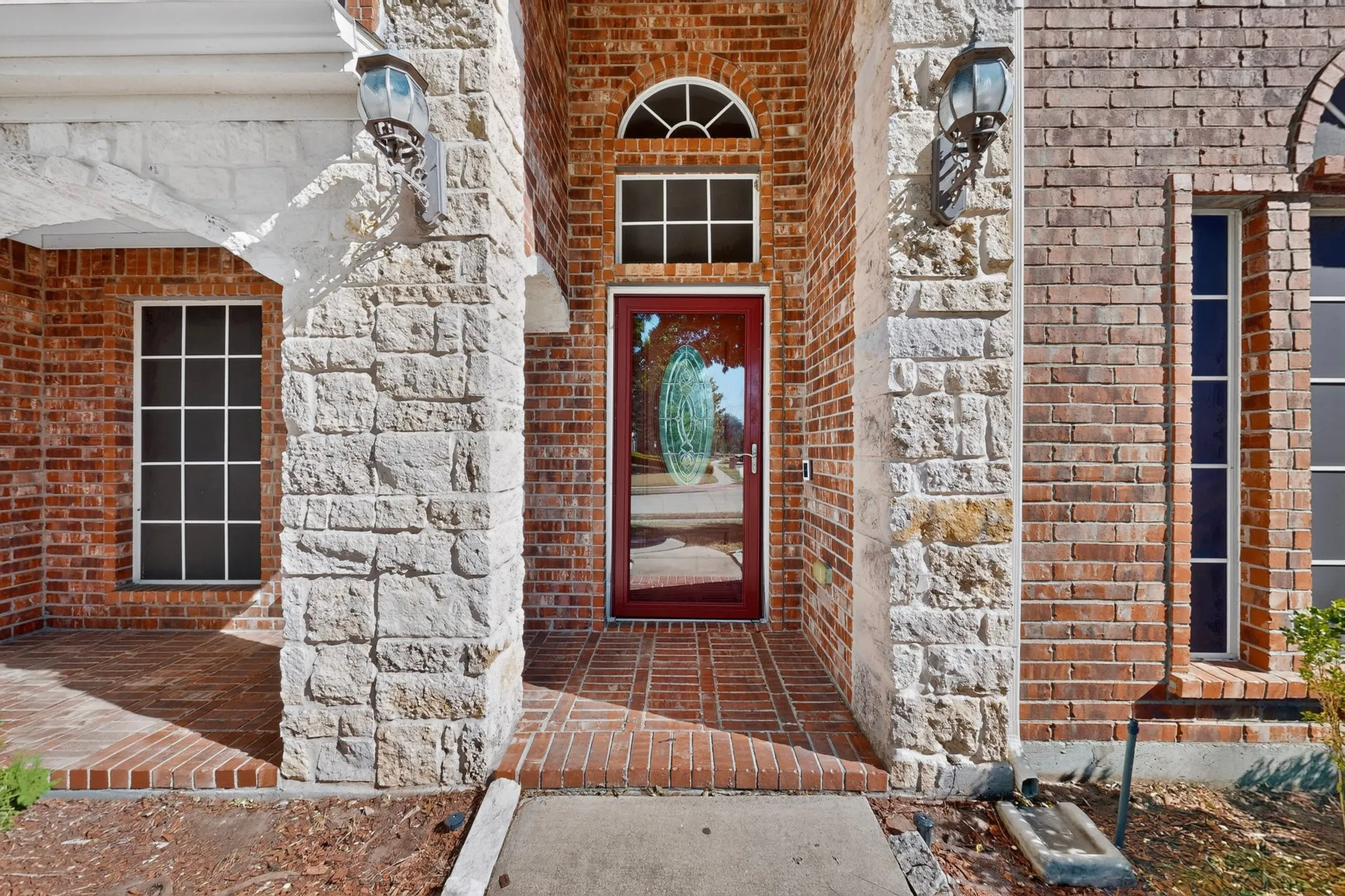 Welcoming covered entry featuring substantial white stone columns, warm brick exterior, and paneled red entry door with sidelights