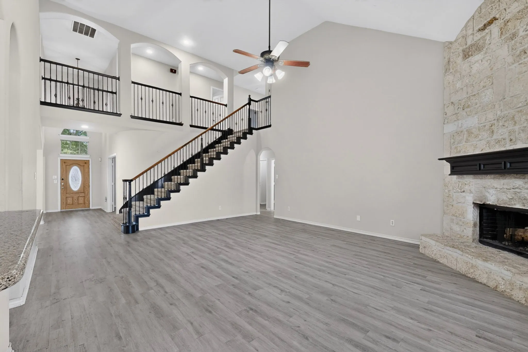 Vaulted ceilings, wood-finish flooring, stone fireplace with dark mantel, wood entry door with oval glass inset, and a wood and wrought iron staircase
