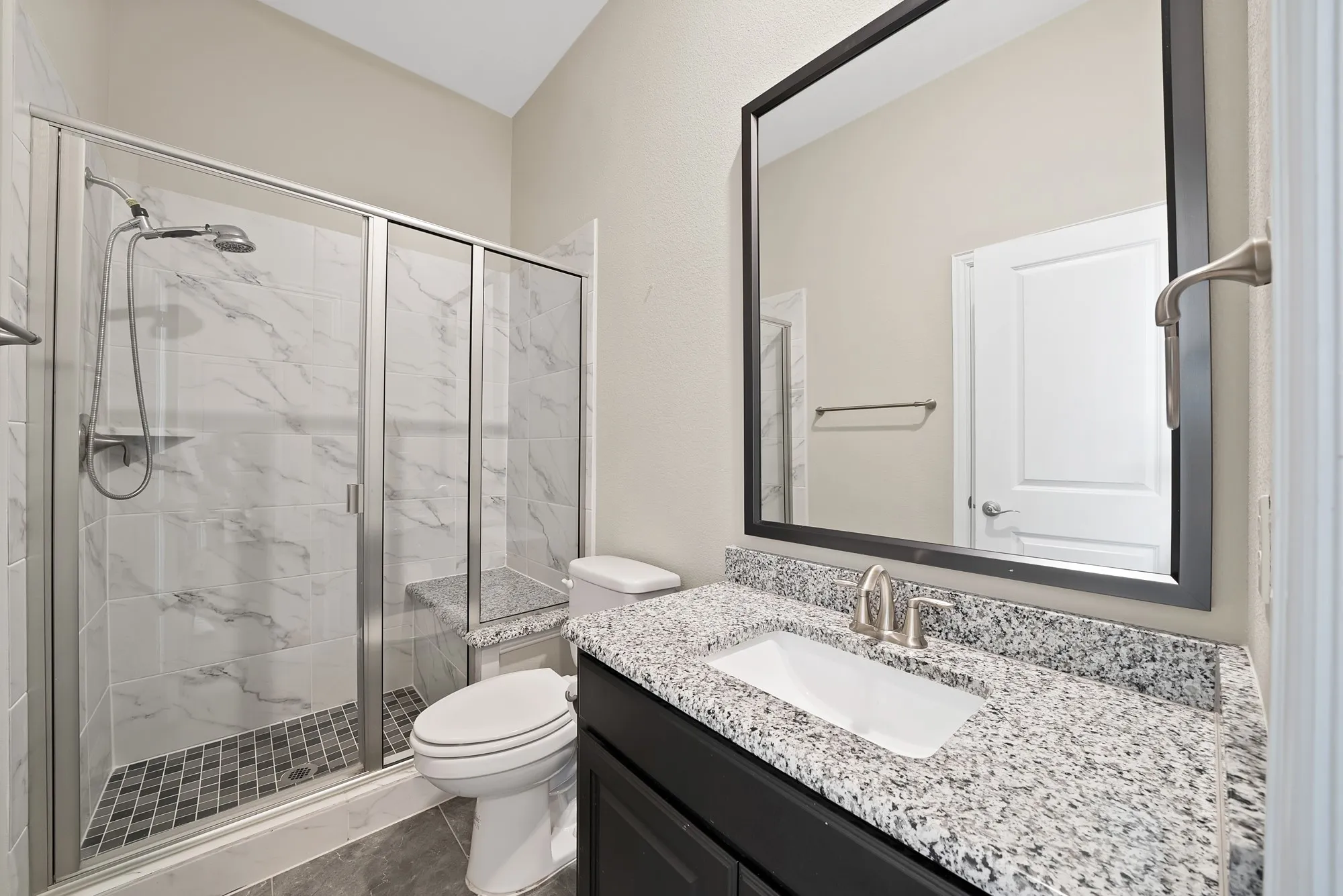 Tiled shower with glass enclosure, integrated shower bench, dark vanity with a light speckled countertop, and a rectangular undermount sink