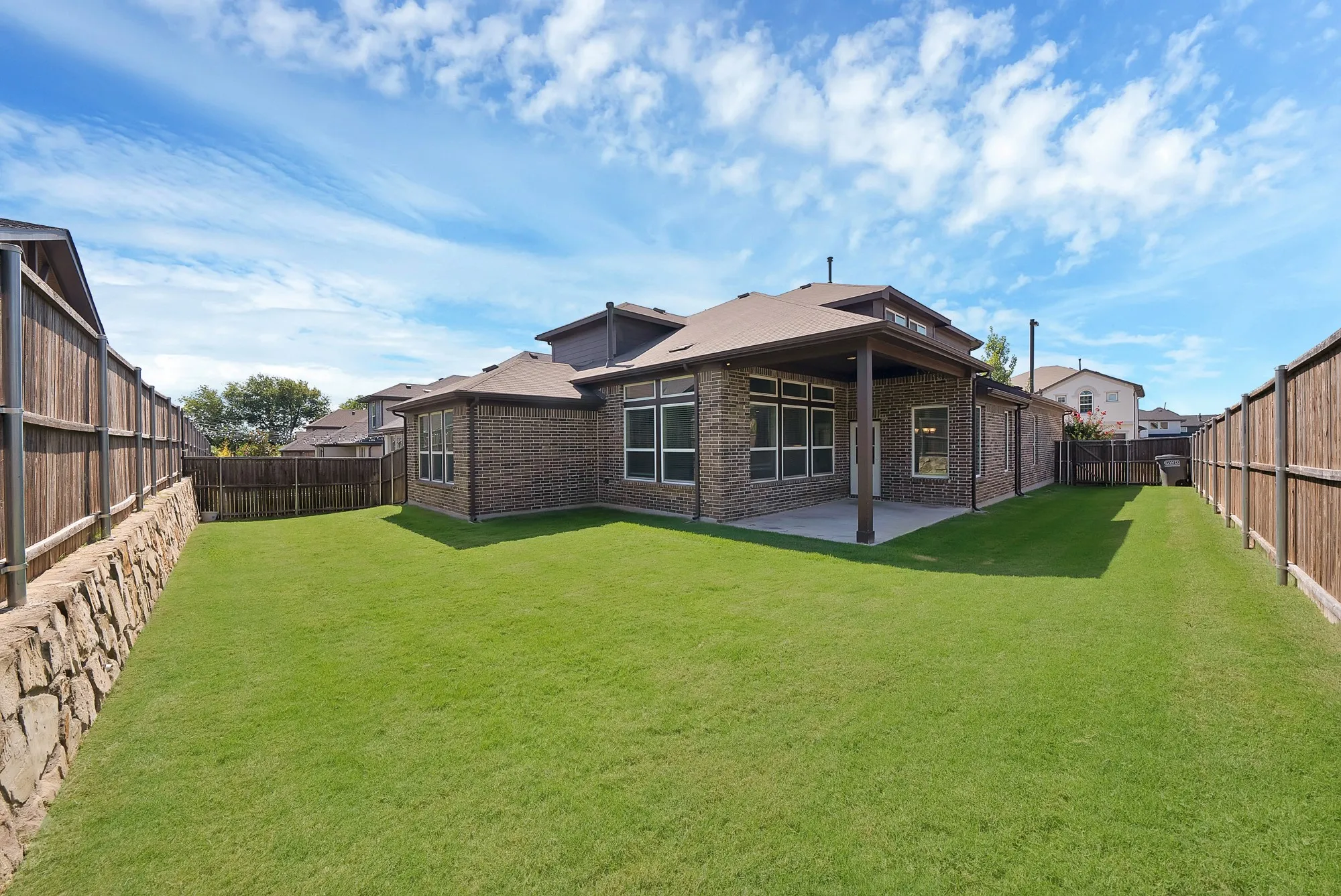 Expansive backyard featuring a manicured lawn, full privacy fencing, a stone retaining wall, and a covered concrete patio