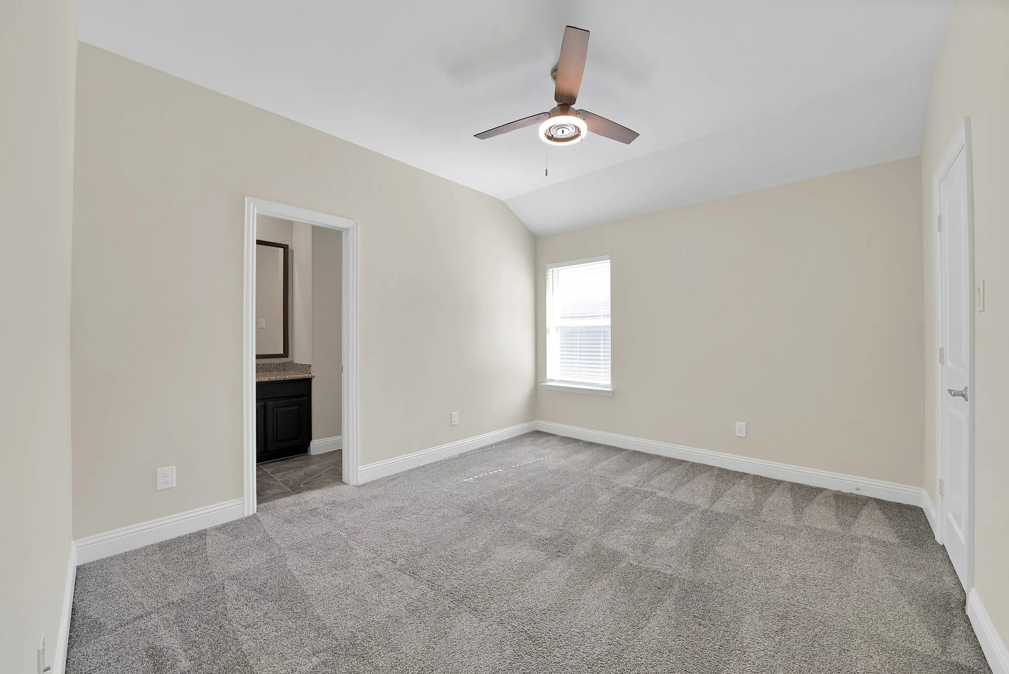 Vaulted ceiling with a contemporary ceiling fan, window with blinds, carpet flooring, and an entryway to a bathroom featuring a vanity with a dark cabinet and a mirror