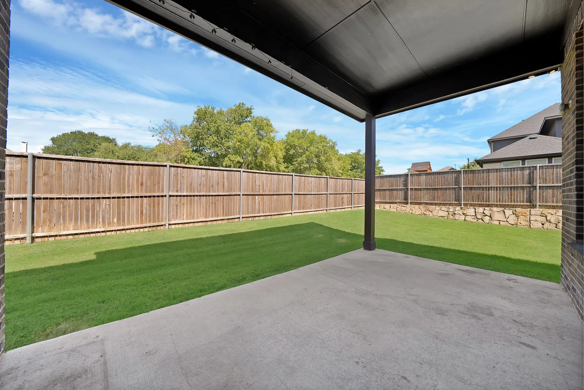 Covered concrete patio opening to a fenced yard with a manicured lawn and a stone retaining wall