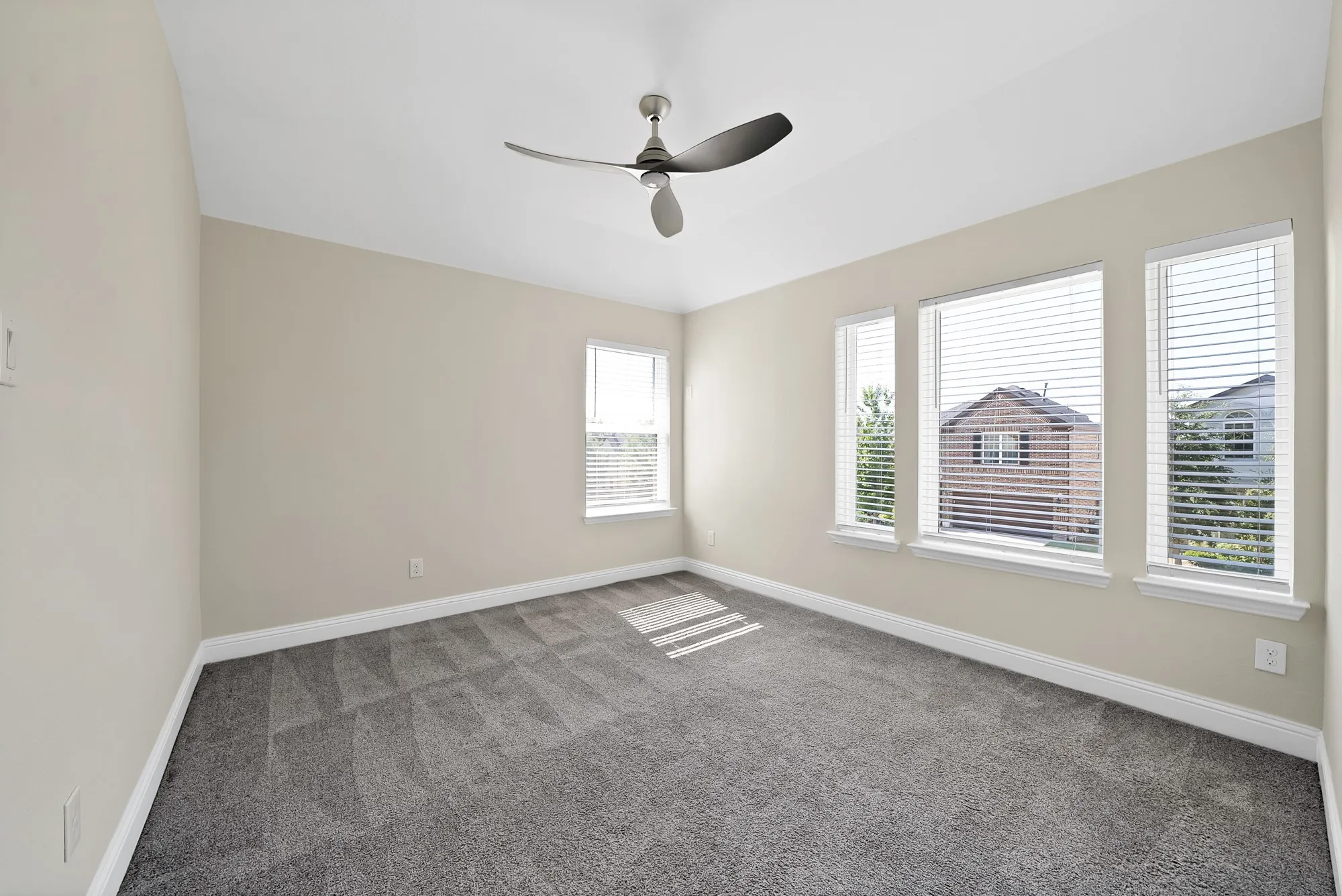 Neutral tone walls, grey patterned carpeting, multiple windows with blinds, and a ceiling fan with integrated lighting