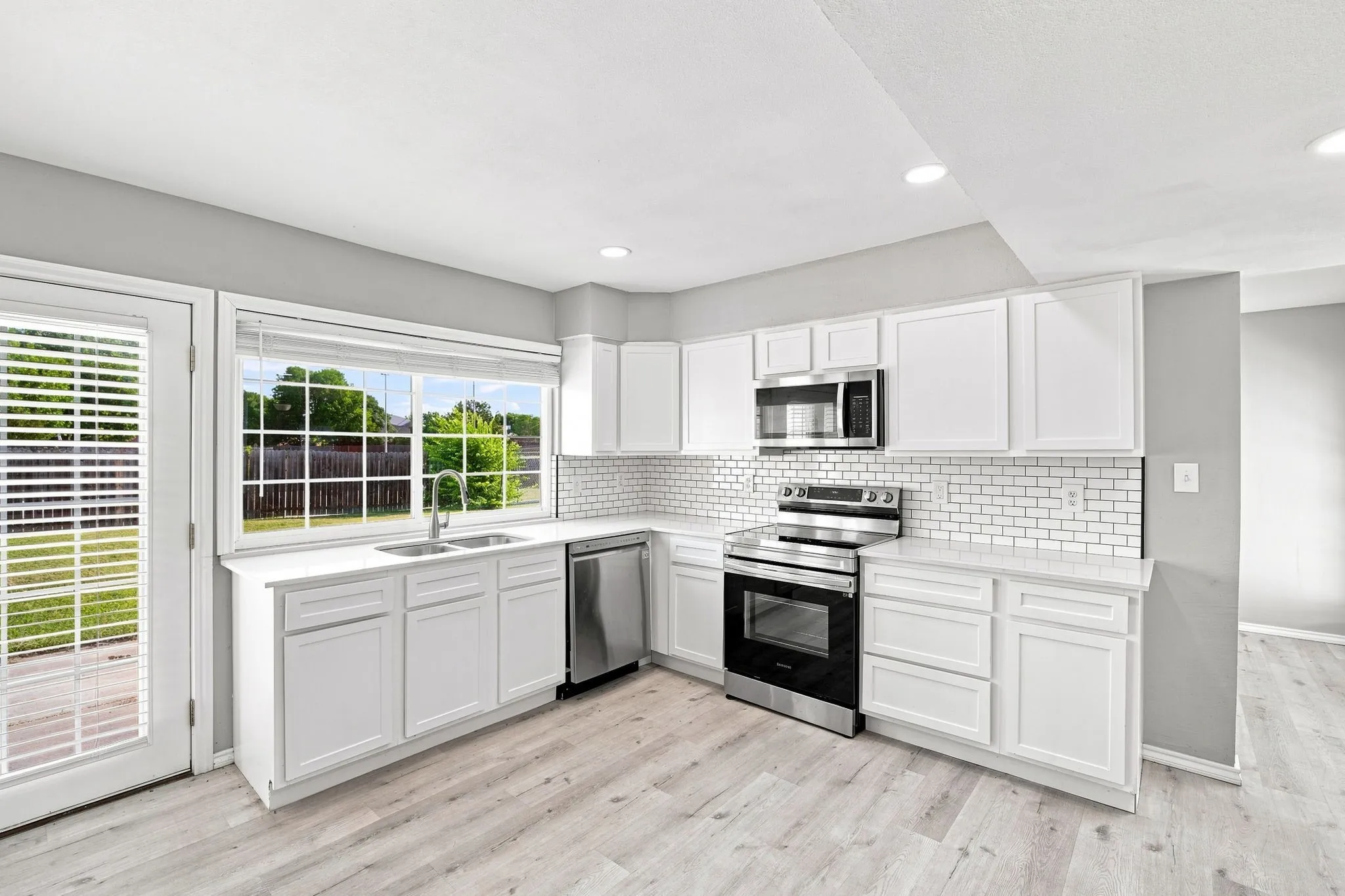 Modern kitchen featuring white cabinetry, stainless steel appliances, subway tile backsplash, wood-finish flooring, and abundant natural light from large windows overlooking the outdoor landscape