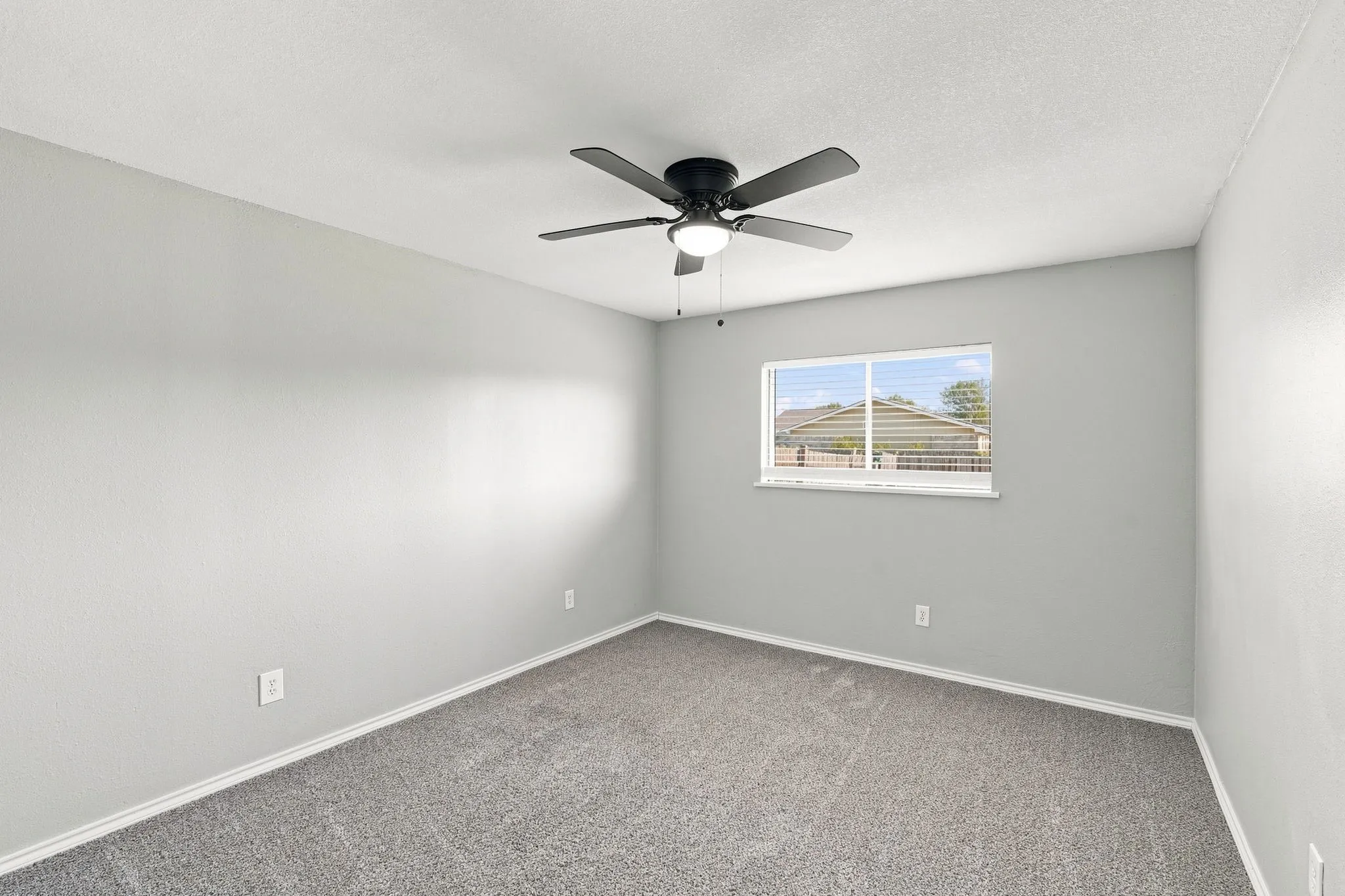 Gray carpeted flooring, light gray painted walls, window with white trim, and a black ceiling fan with integrated lighting