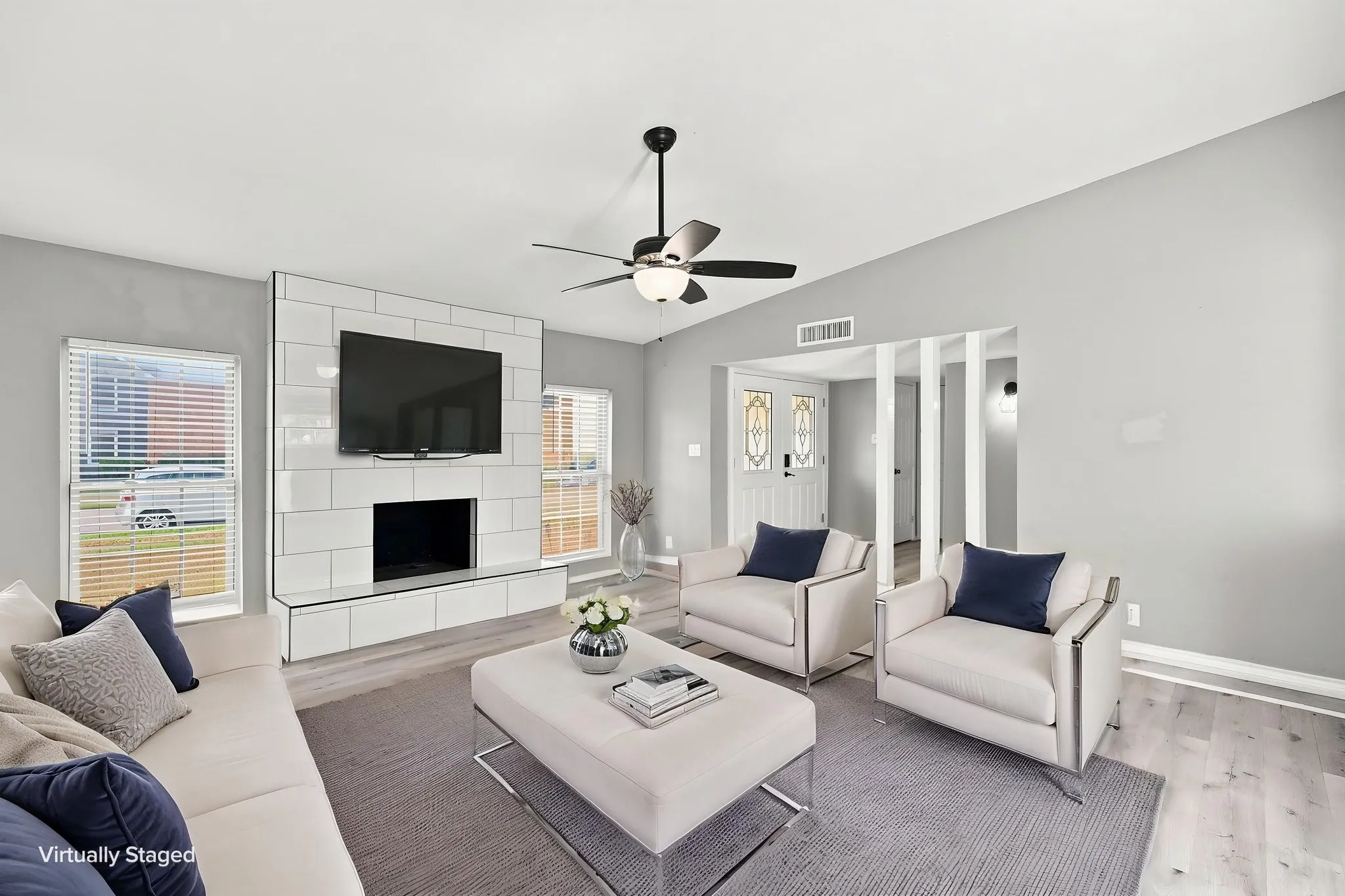Sleek tiled fireplace with integrated hearth, ceiling fan with illumination, light wood-finish flooring, and two front-facing windows