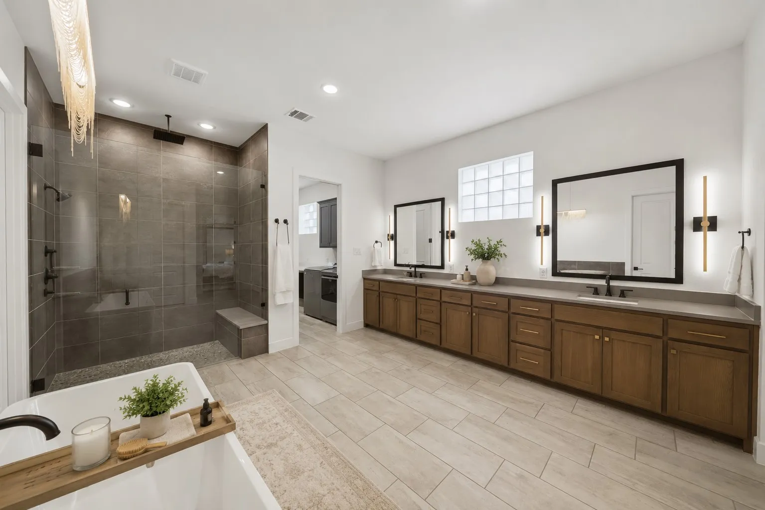 Spacious bathroom featuring a built-in shower bench, dual vanities with dark cabinetry, light-toned countertops and direct access to laundry room.