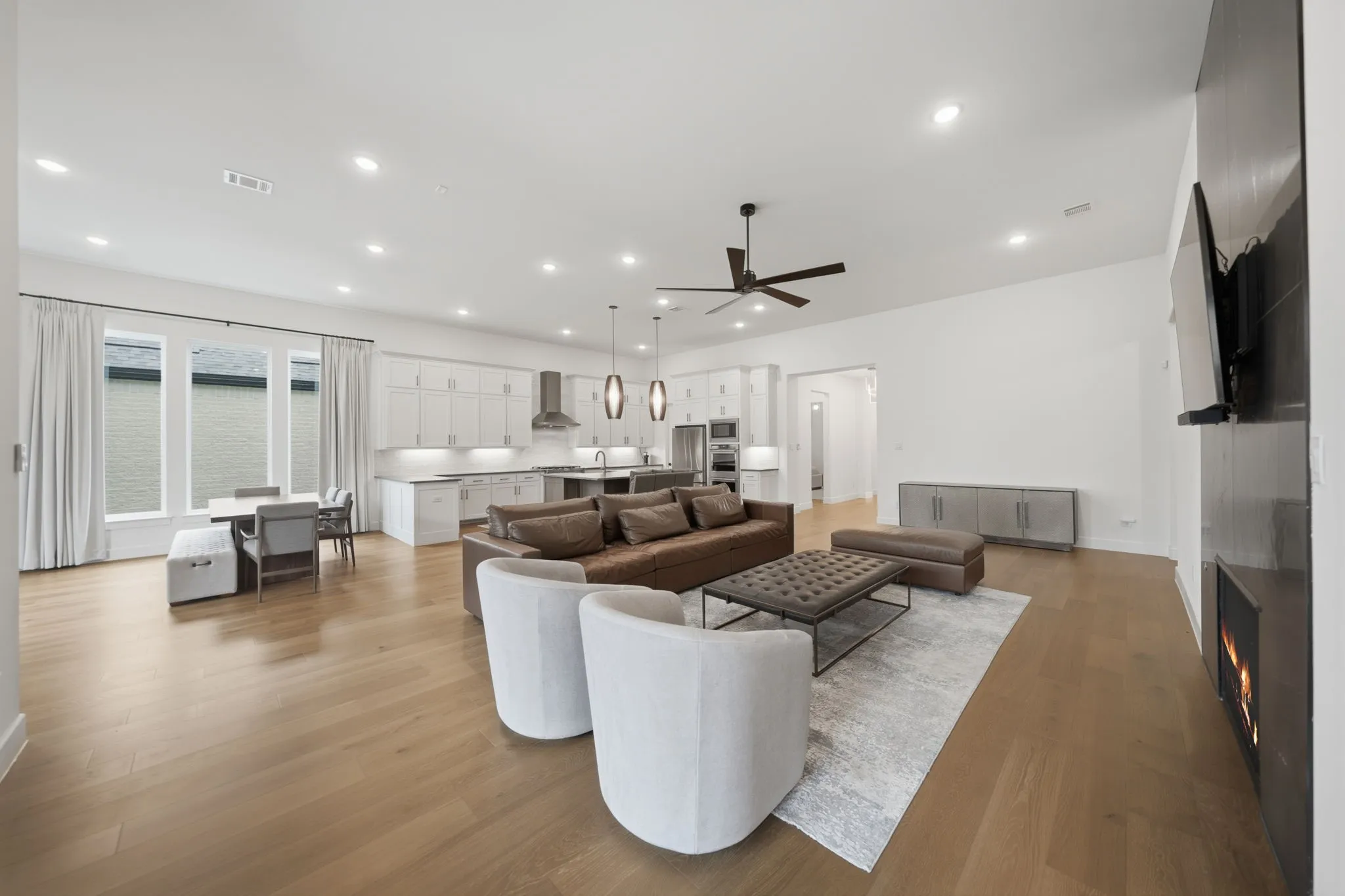 Expansive open-concept space featuring wood-finish flooring, a contemporary kitchen with white cabinetry and stainless steel appliances, a built-in electric fireplace, and recessed lighting throughout