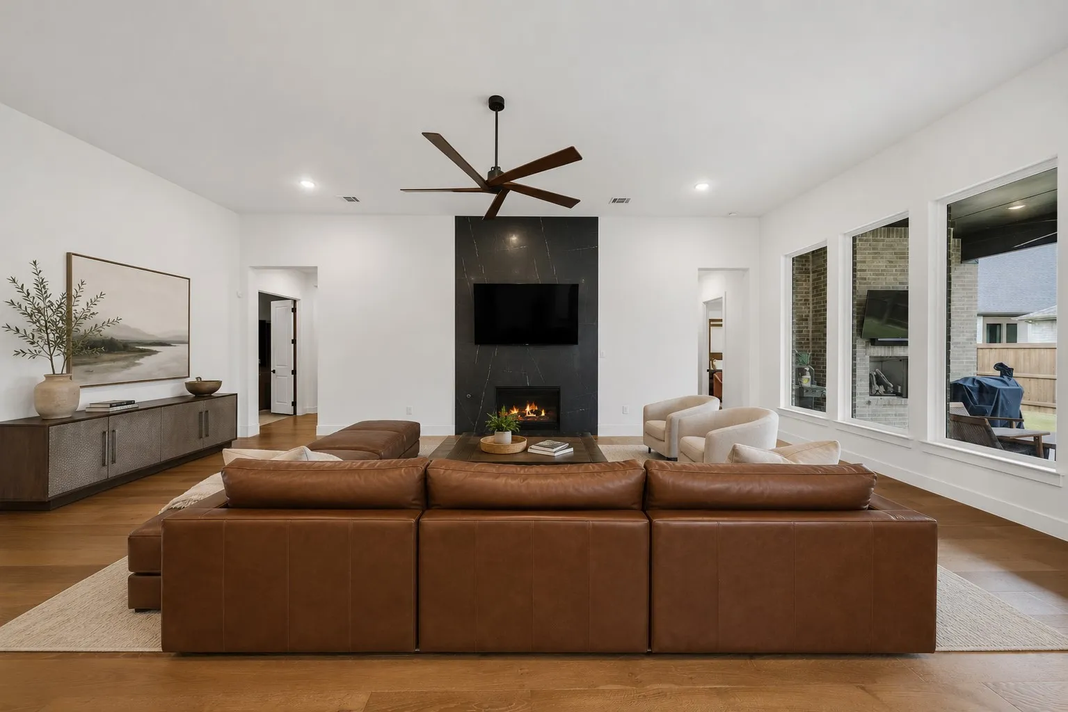 Spacious living area featuring wood-finish flooring, a contemporary fireplace with a dark stone surround, a ceiling fan, and multiple large windows