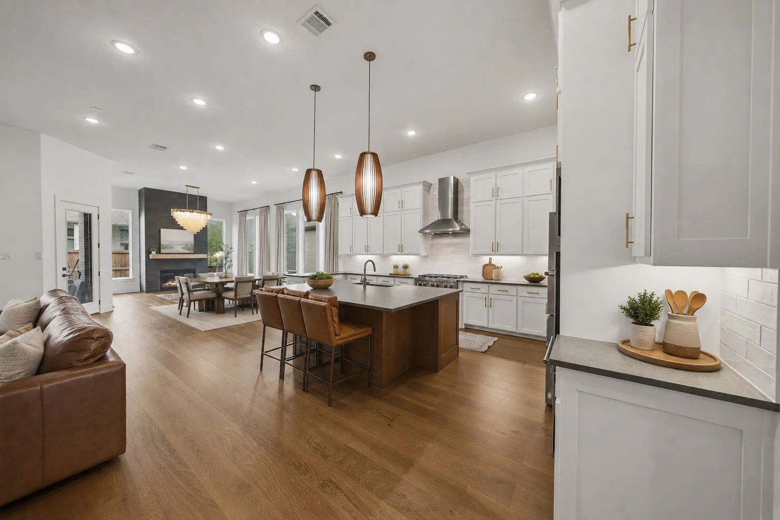Open-concept living area with wood-finish flooring, a kitchen island with pendant lighting, white cabinetry, stainless steel appliances, and a fireplace with a dark stone surround