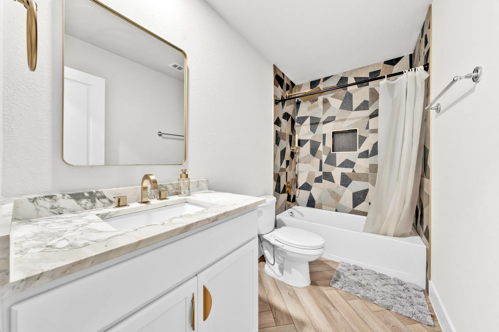 Vanity with a single undermount sink and brass-finish faucet, framed mirror, built-in bathtub with shower, geometric patterned wall tile, and wood-finish flooring