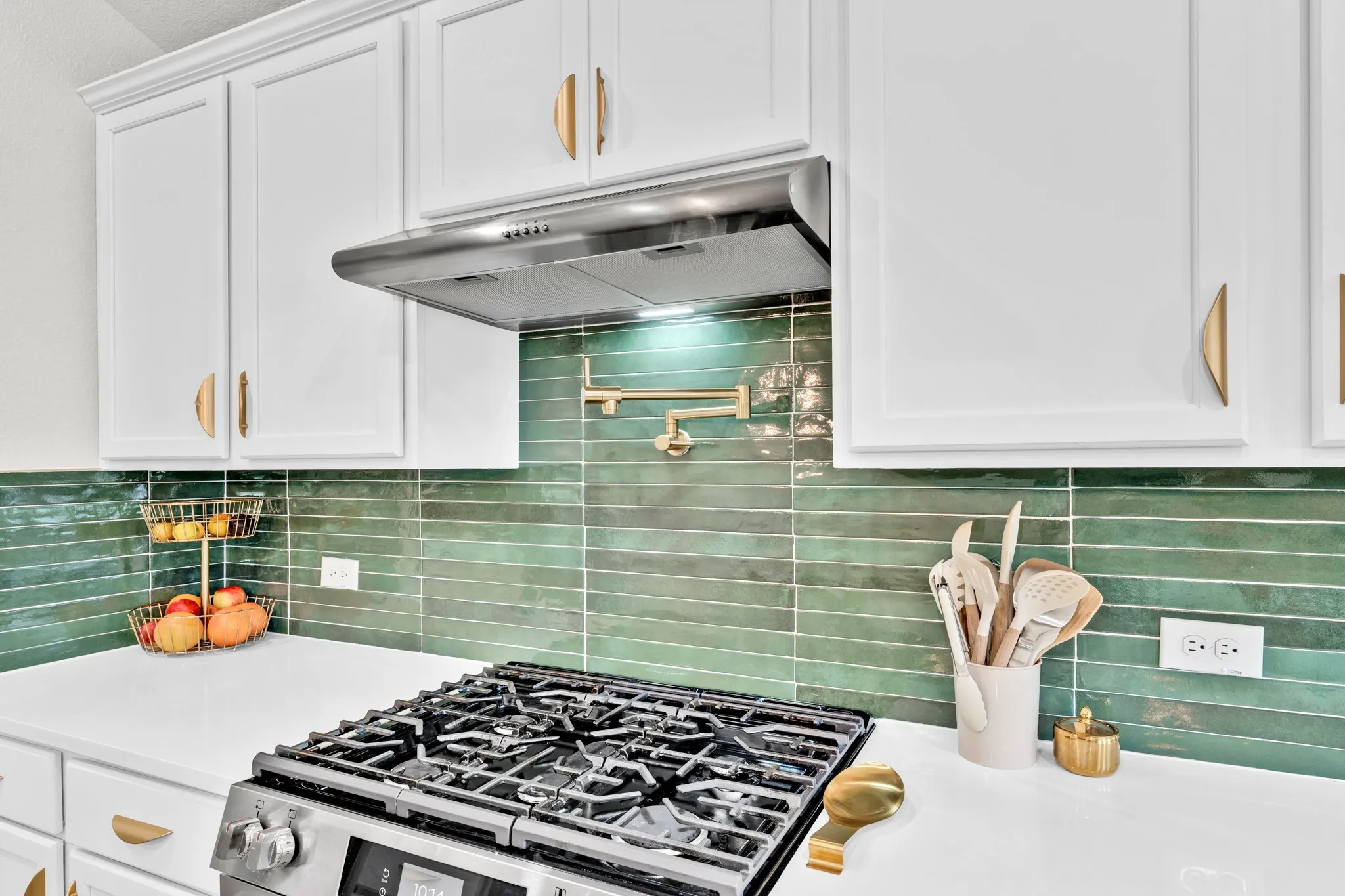 White cabinetry with gold-tone hardware, stainless steel range with overhead hood, green subway tile backsplash, white countertops, and a wall-mounted pot filler