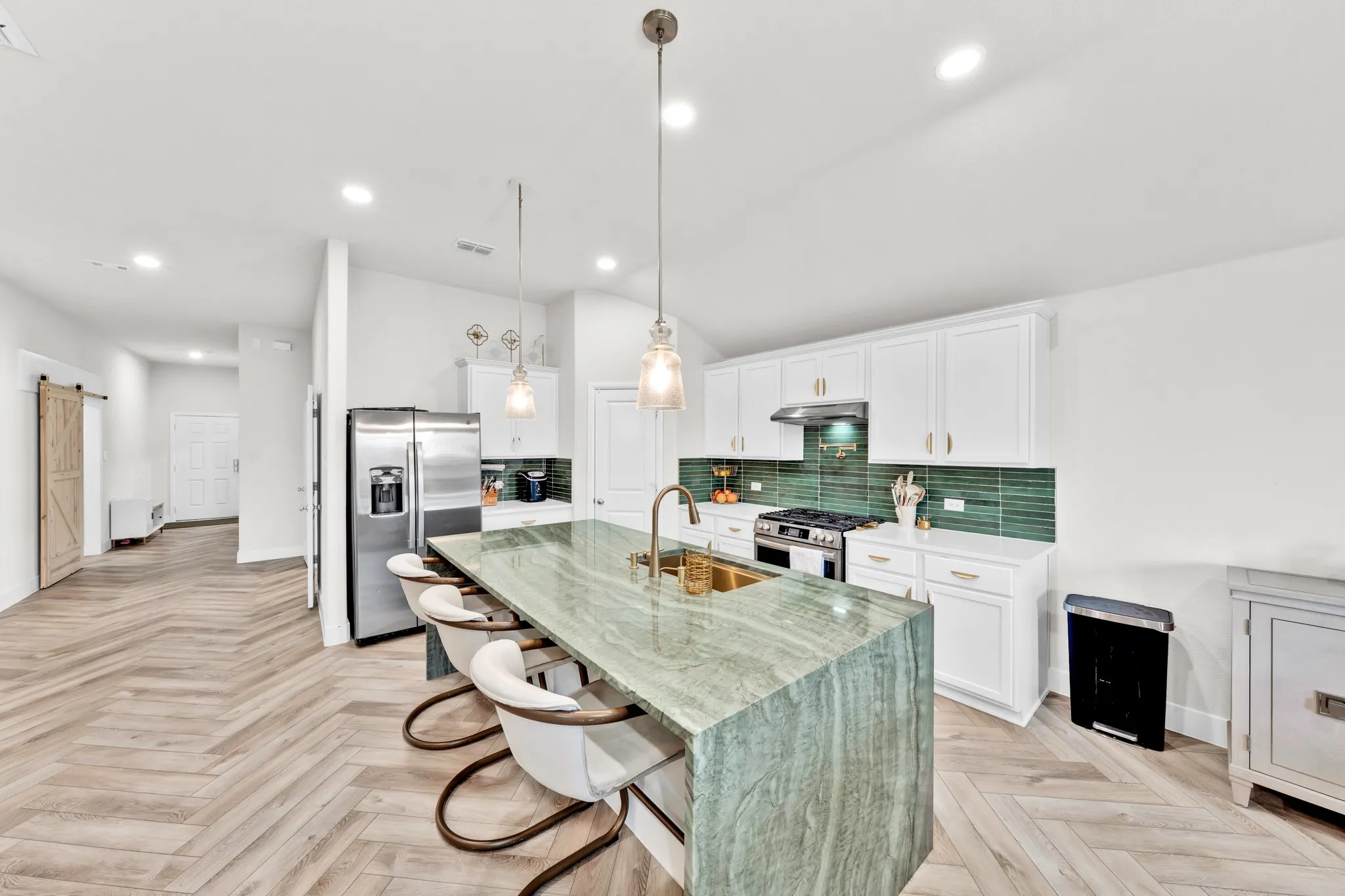 Spacious kitchen featuring a waterfall island, stainless steel appliances, white cabinetry with gold-tone hardware, and a vibrant green tile backsplash