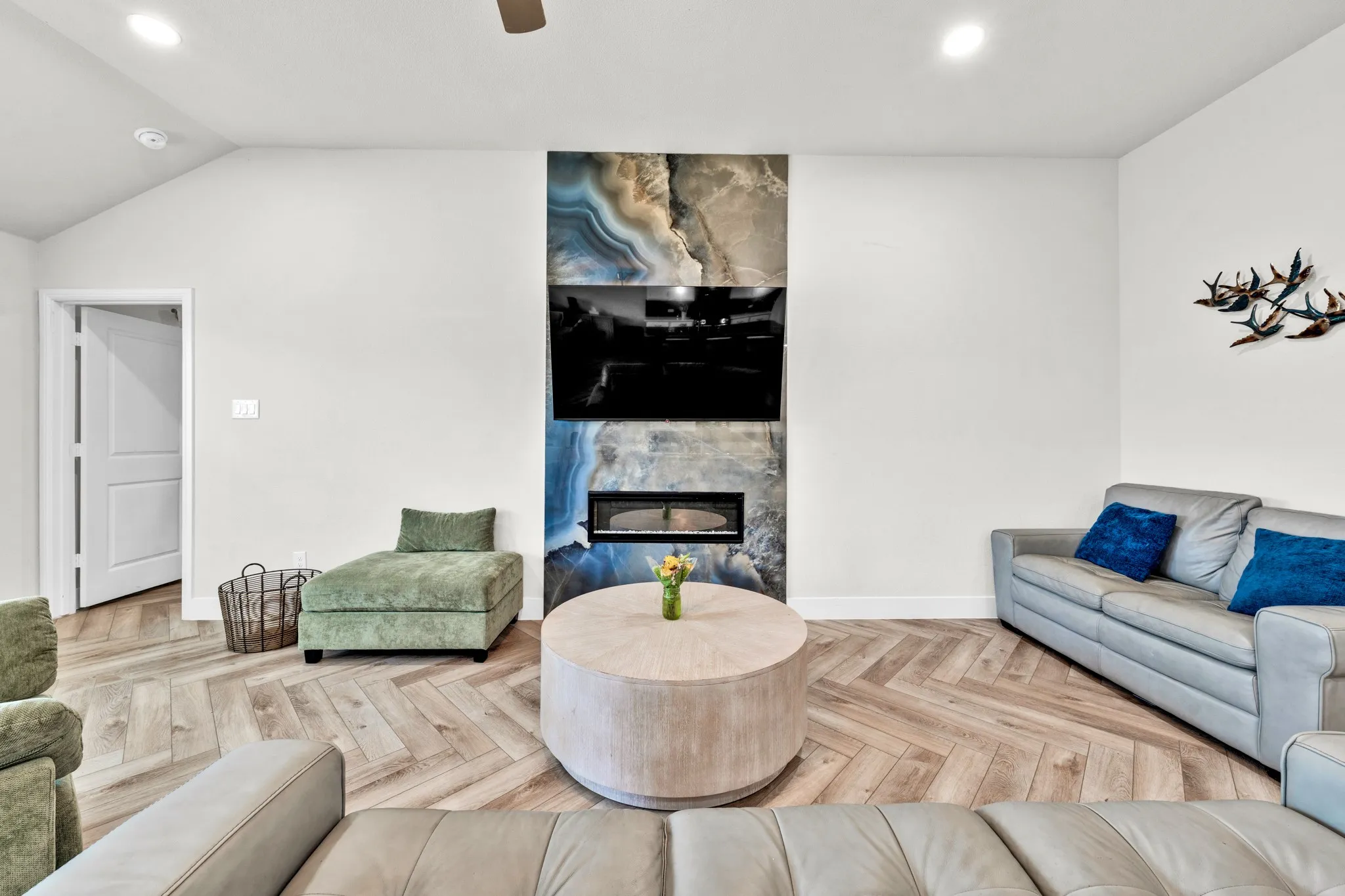 Vaulted ceiling with recessed lighting, wood-finish flooring in a herringbone pattern, built-in electric fireplace with stone surround, and an integrated ceiling fan