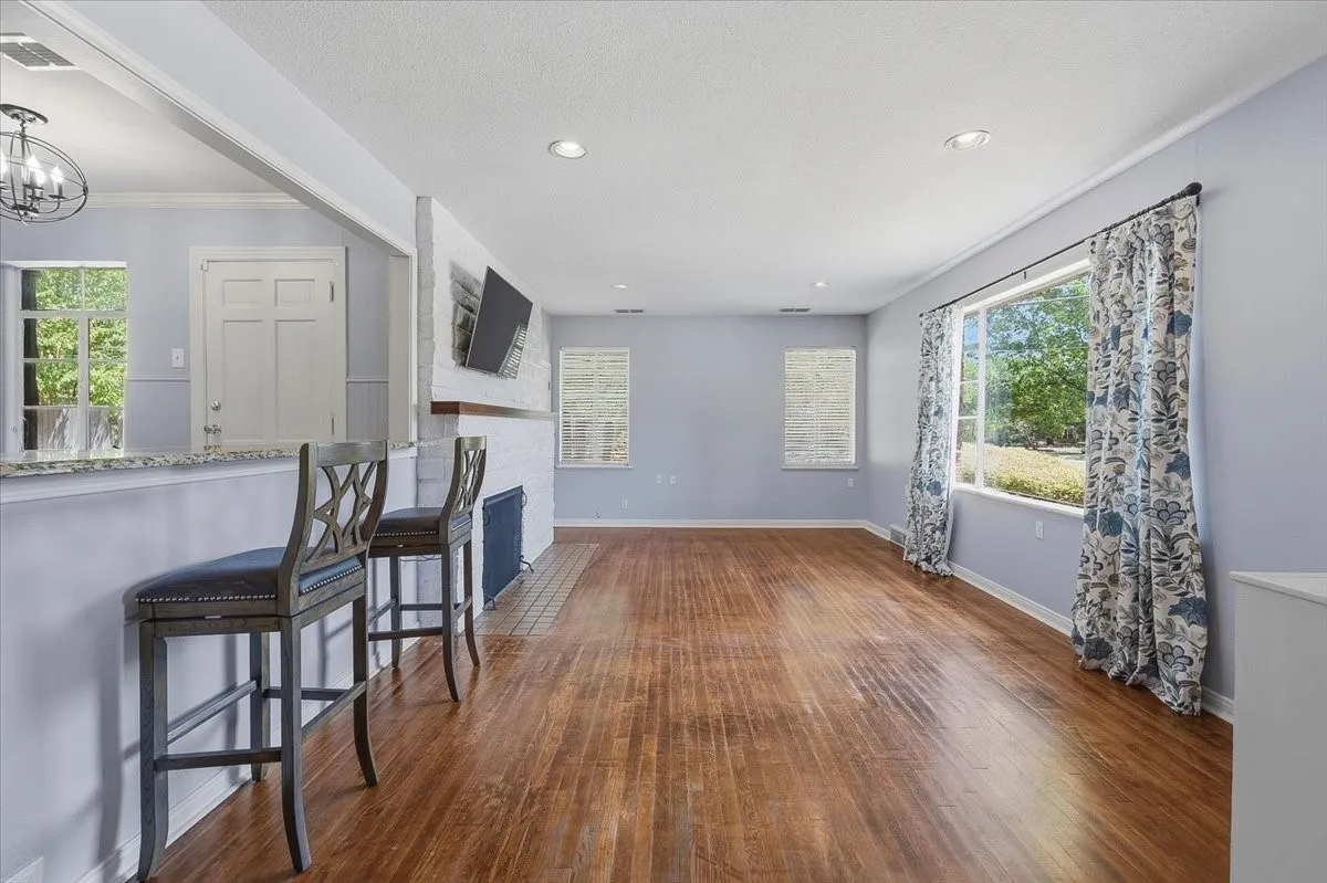 Spacious living area featuring rich hardwood flooring, a white brick fireplace with black surround, wall-mounted flat-screen television, recessed ceiling lighting, and an open layout connecting to the kitchen with bar seating