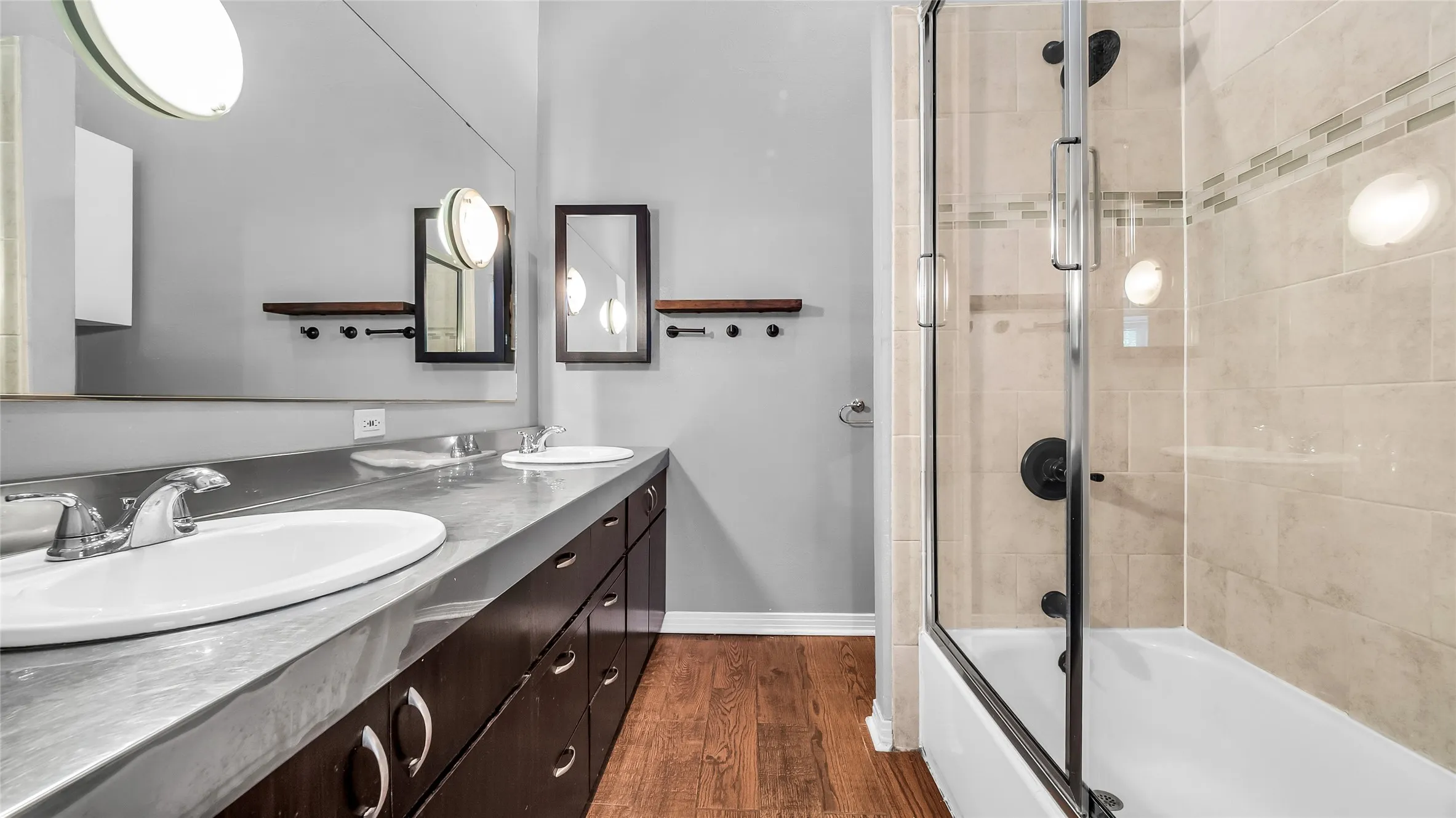 Dual vanity with integrated sinks, extensive dark wood-finish cabinetry, a large wall-mounted mirror, wood-finish flooring, and a tiled shower with a glass enclosure