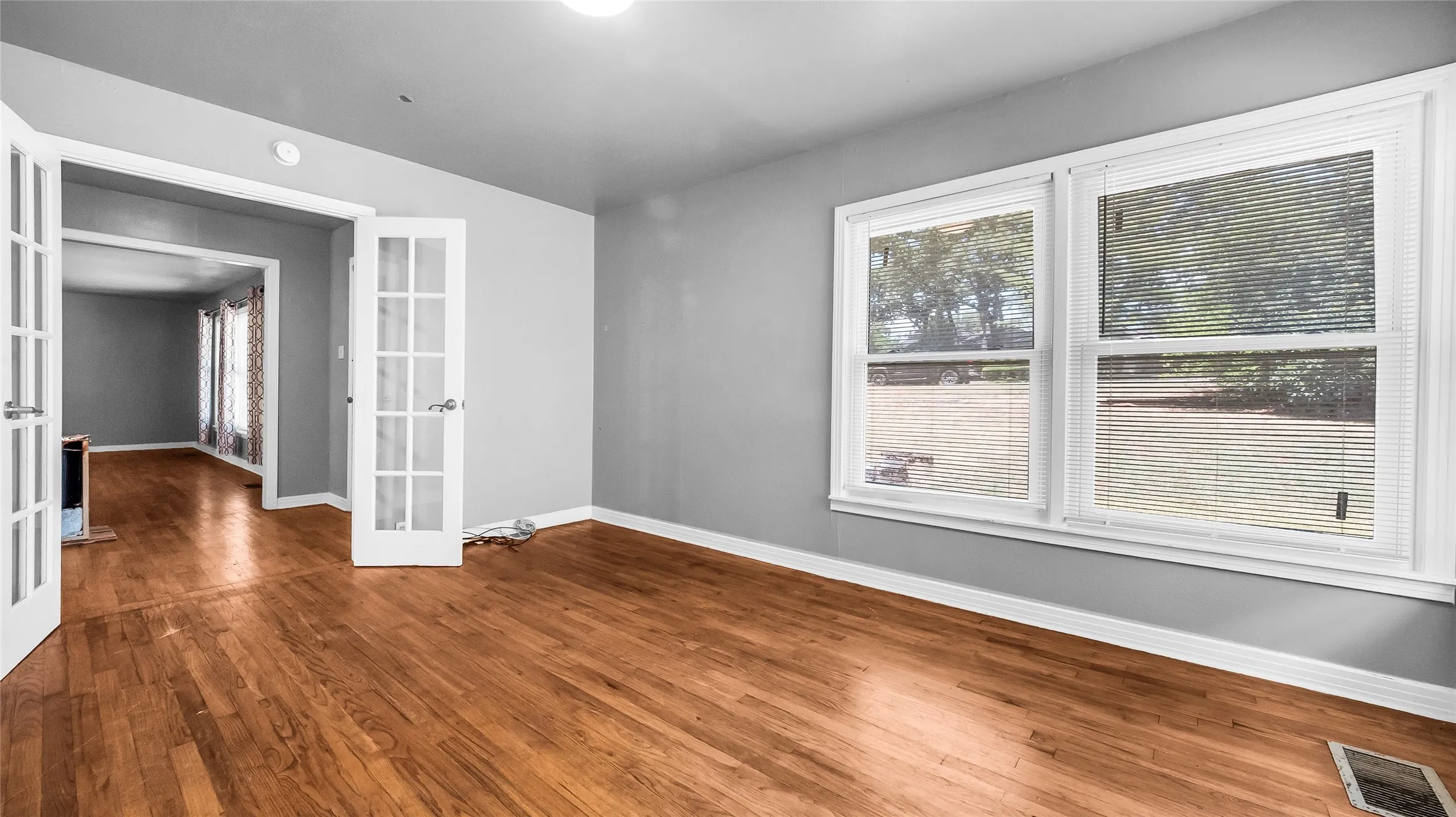 Wood-finish flooring throughout, French doors with clear glass panes, and two large windows featuring white blinds