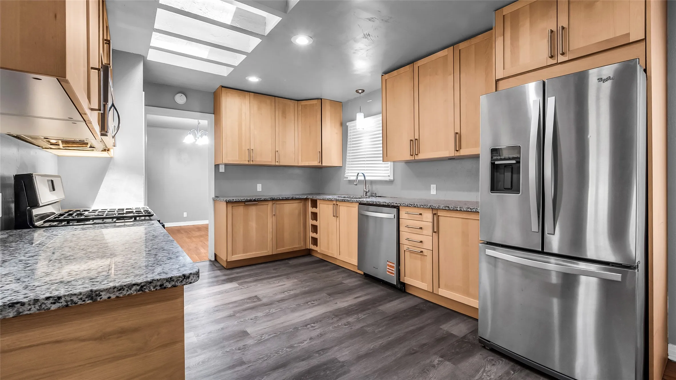 Kitchen featuring light wood cabinetry, granite-style countertops, stainless steel appliances, wood-finish flooring, and a skylight