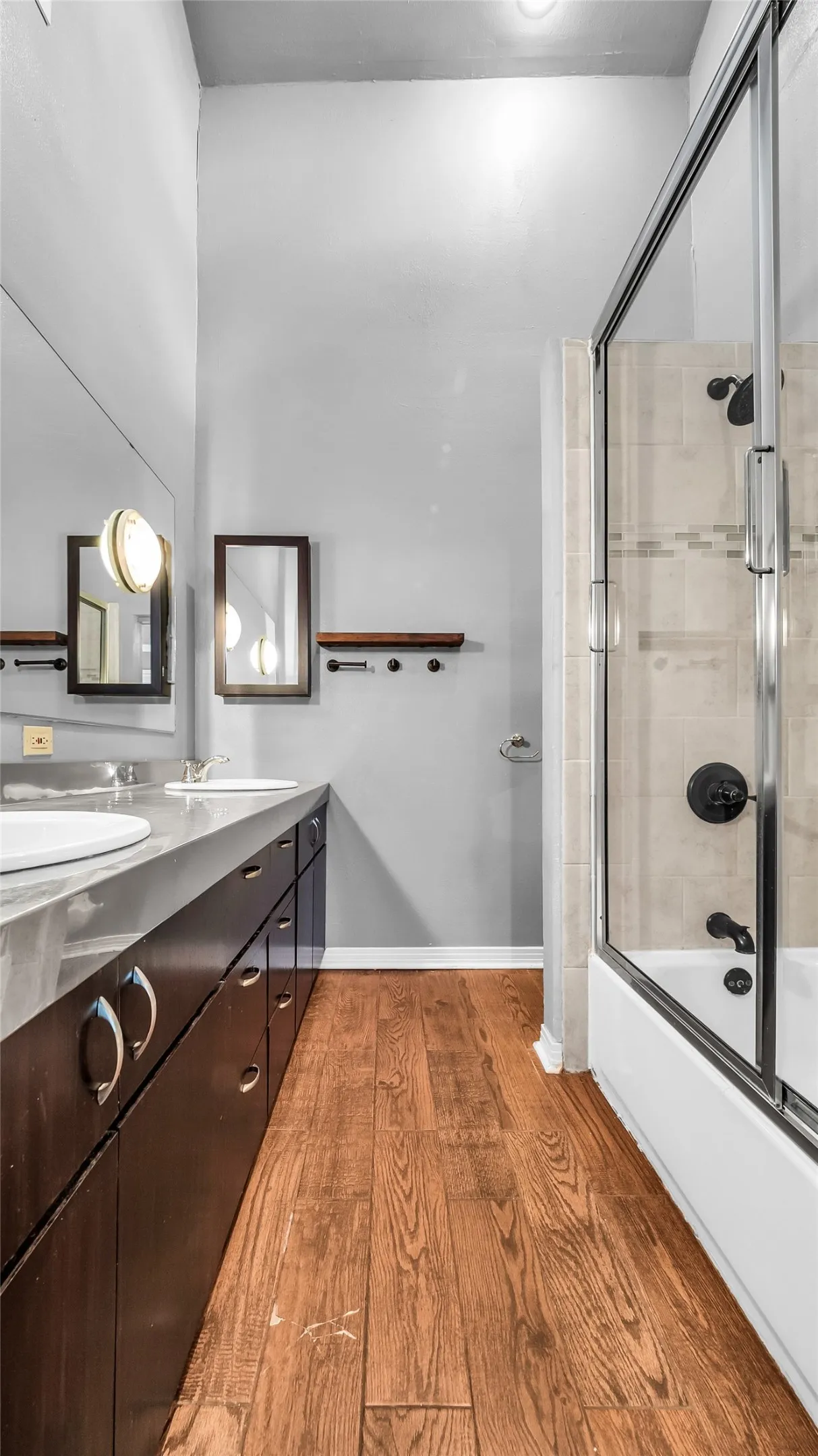 Wood-finish flooring with an extensive double vanity featuring integrated sinks, dark cabinetry, and a shower-over-tub combination with a glass enclosure